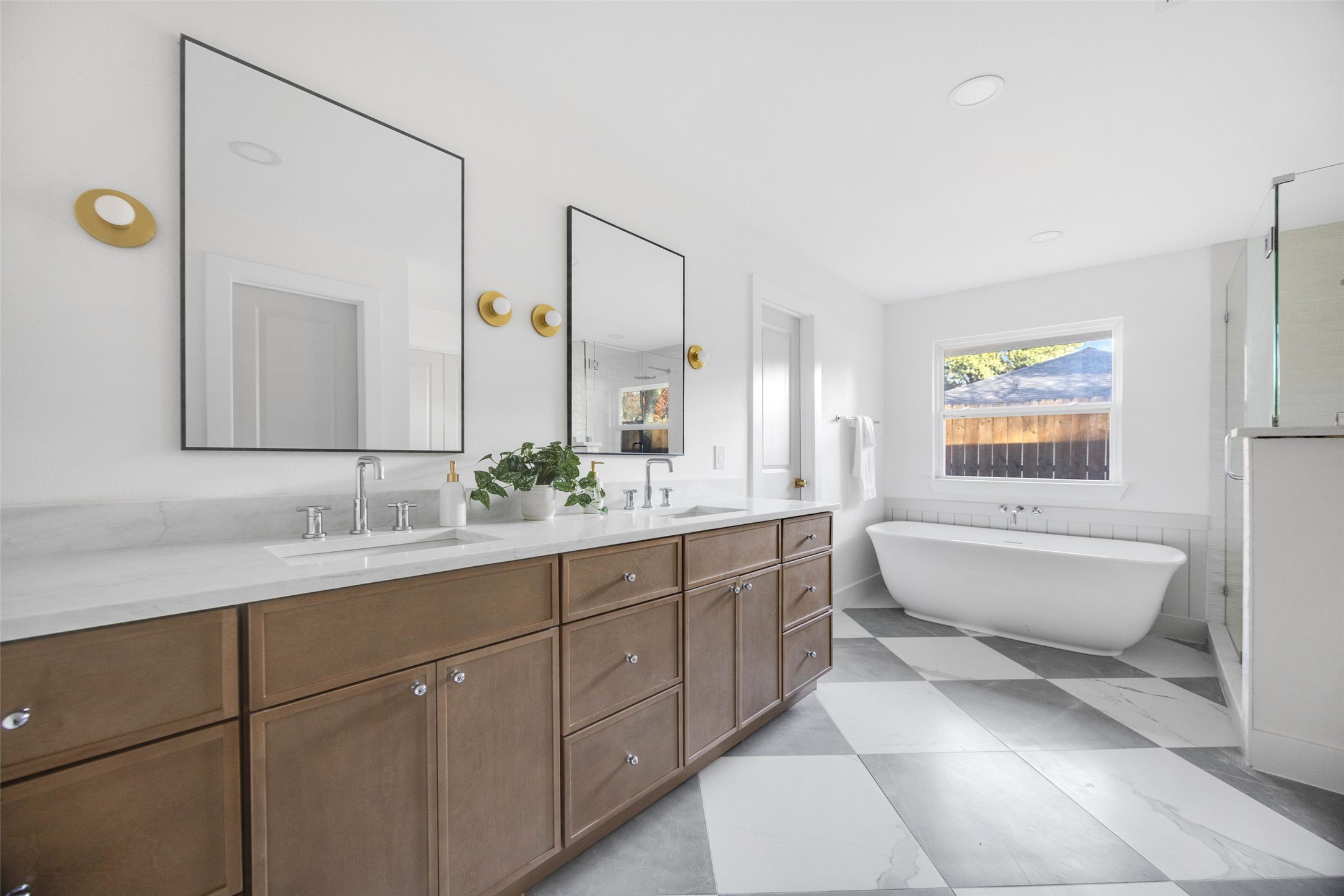 Bathroom with double-sink wood vanity, freestanding tub beneath a window, and glass shower.