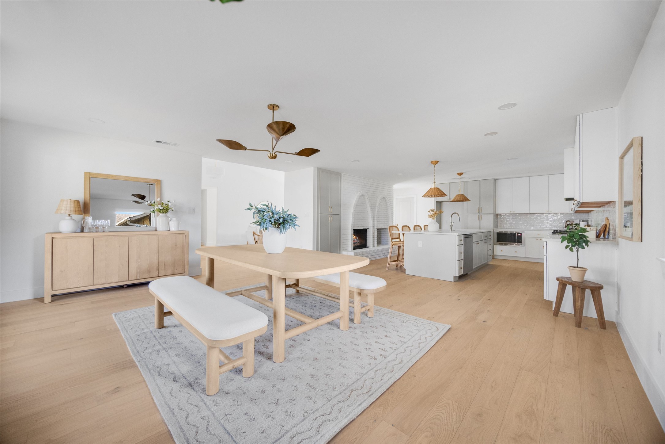 Open-plan dining room and kitchen with a light-wood table, central island, and white brick fireplace.
