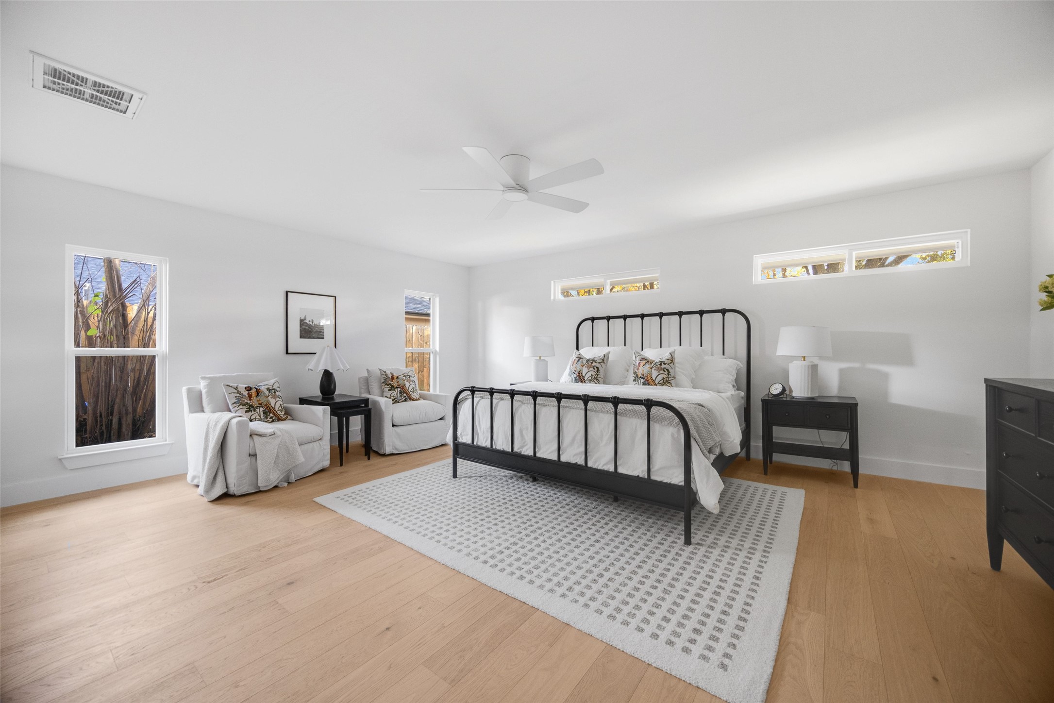 Staged bedroom with a black metal bed, white bedding, and two chairs beside tall windows.
