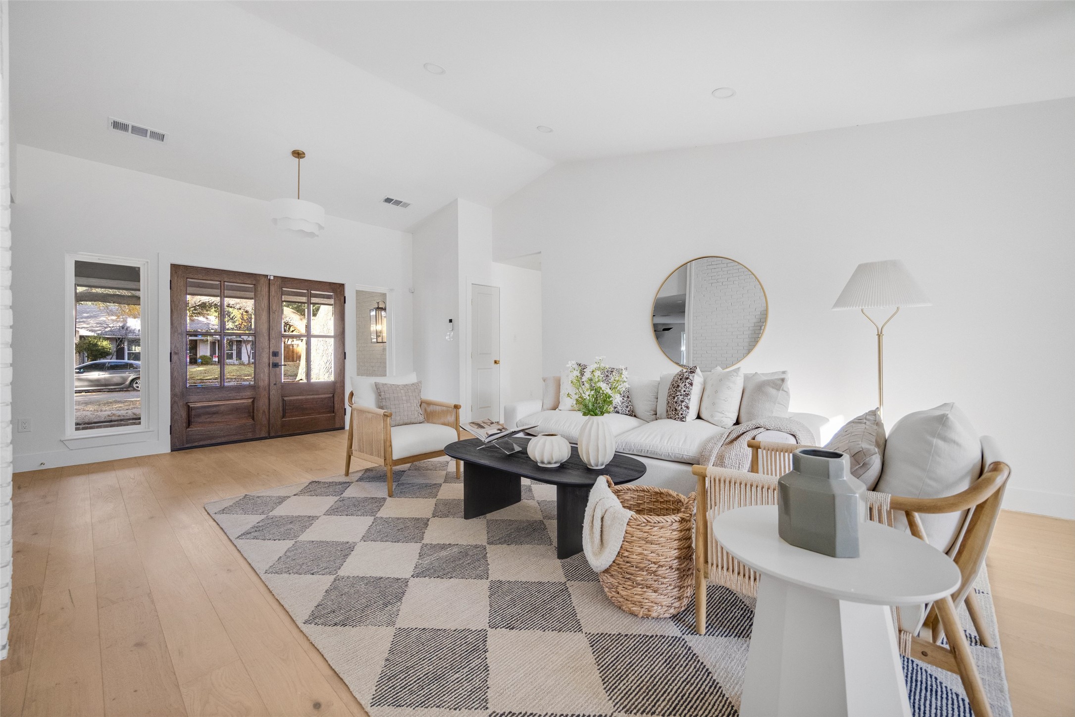 Staged living room with vaulted ceiling, wood-and-glass entry doors, white seating, and a large checkered rug.