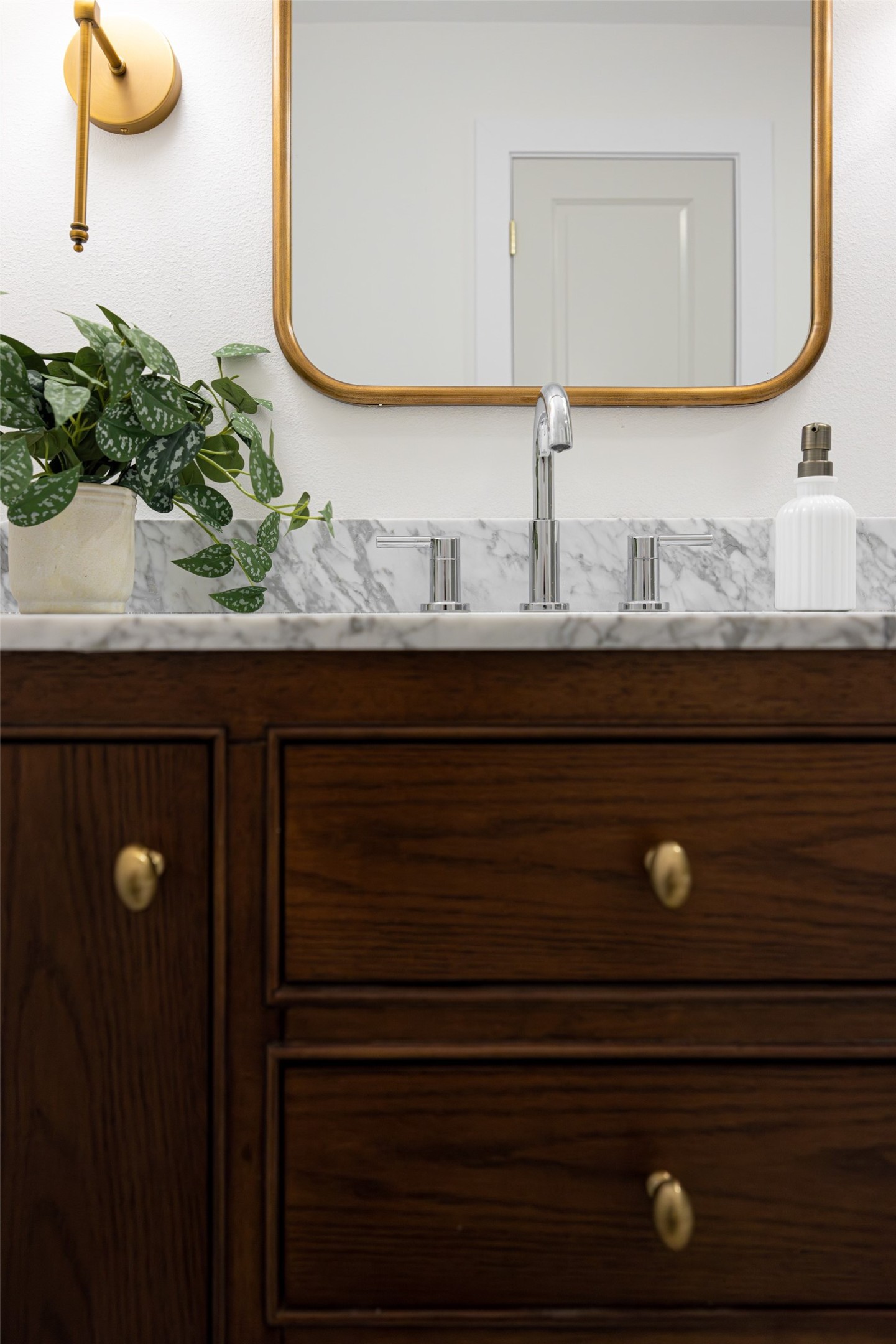 Bathroom vanity with dark wood cabinets, marble surfaces, a brass-framed mirror, and a potted plant.