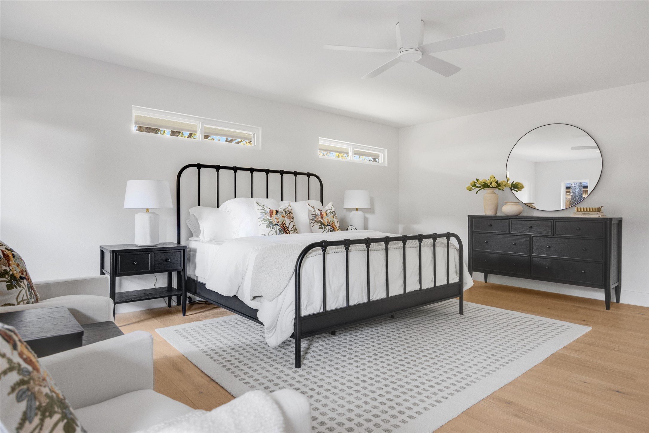 Staged bedroom with a black metal bed, white bedding, high windows, and a dark dresser with round mirror.