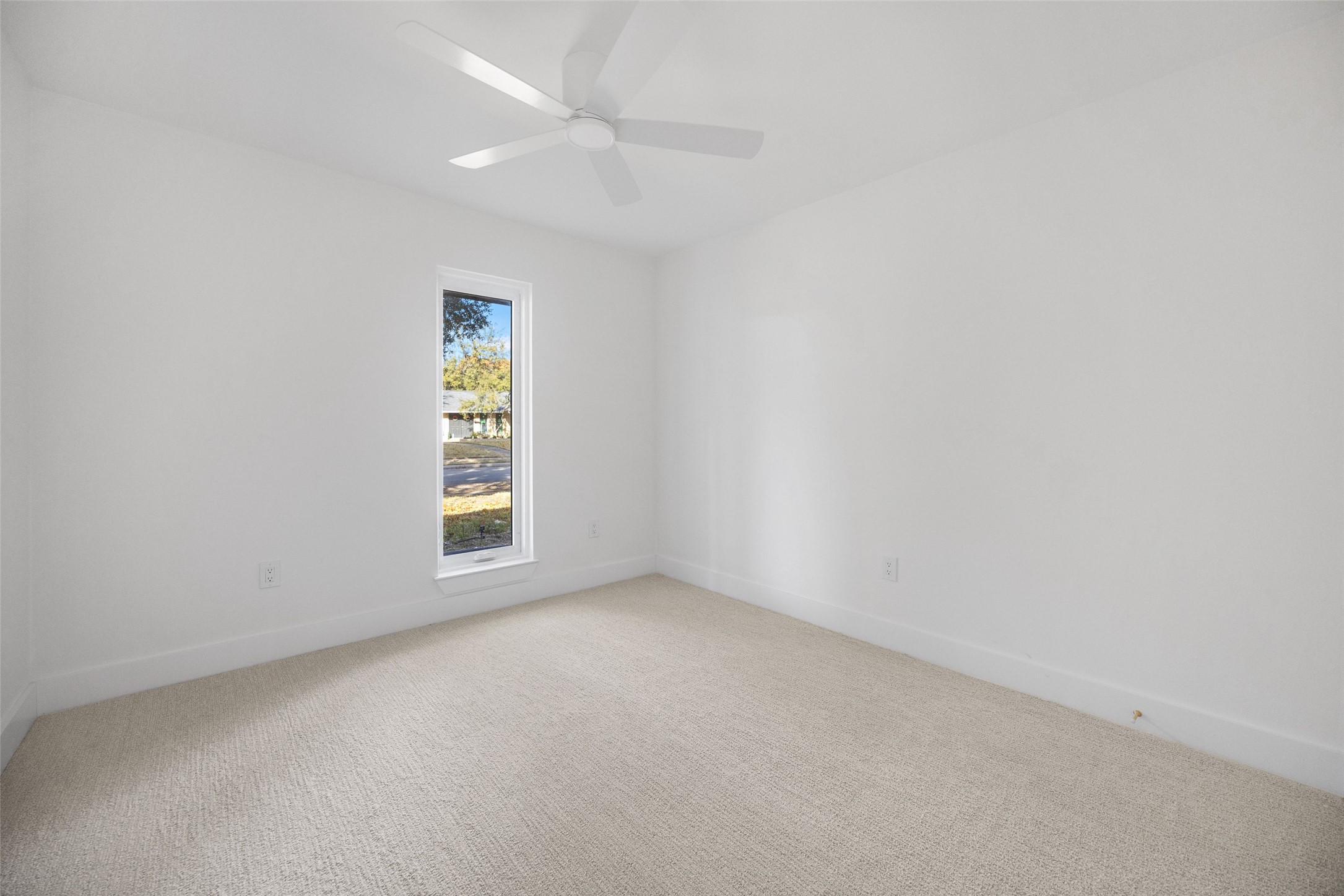 Empty bedroom with beige carpet, a narrow window, and a white ceiling fan.