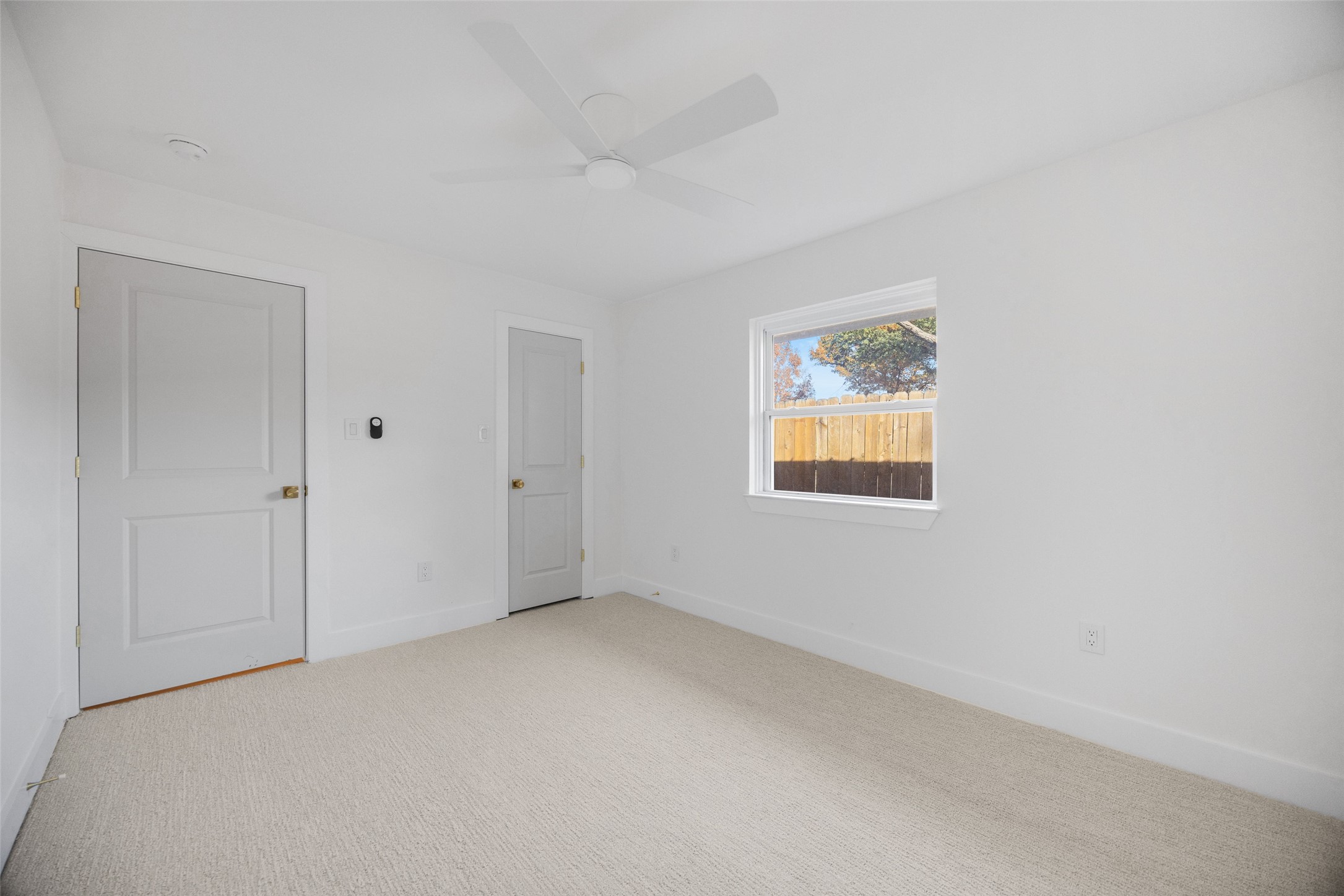 Empty carpeted bedroom with a ceiling fan and a window overlooking a wooden fence.