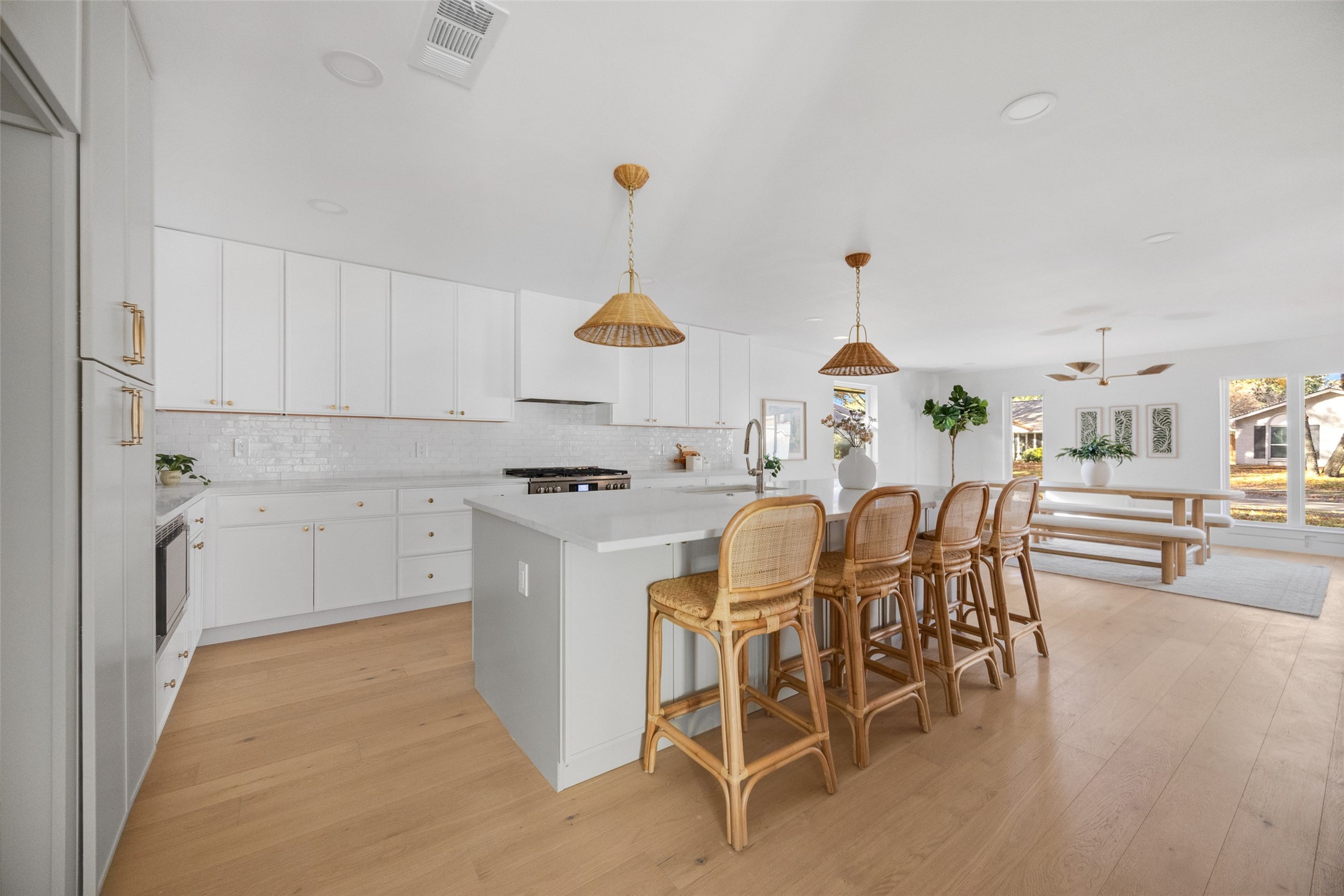 Open kitchen with a large island and woven bar stools beside a dining area with wide windows.