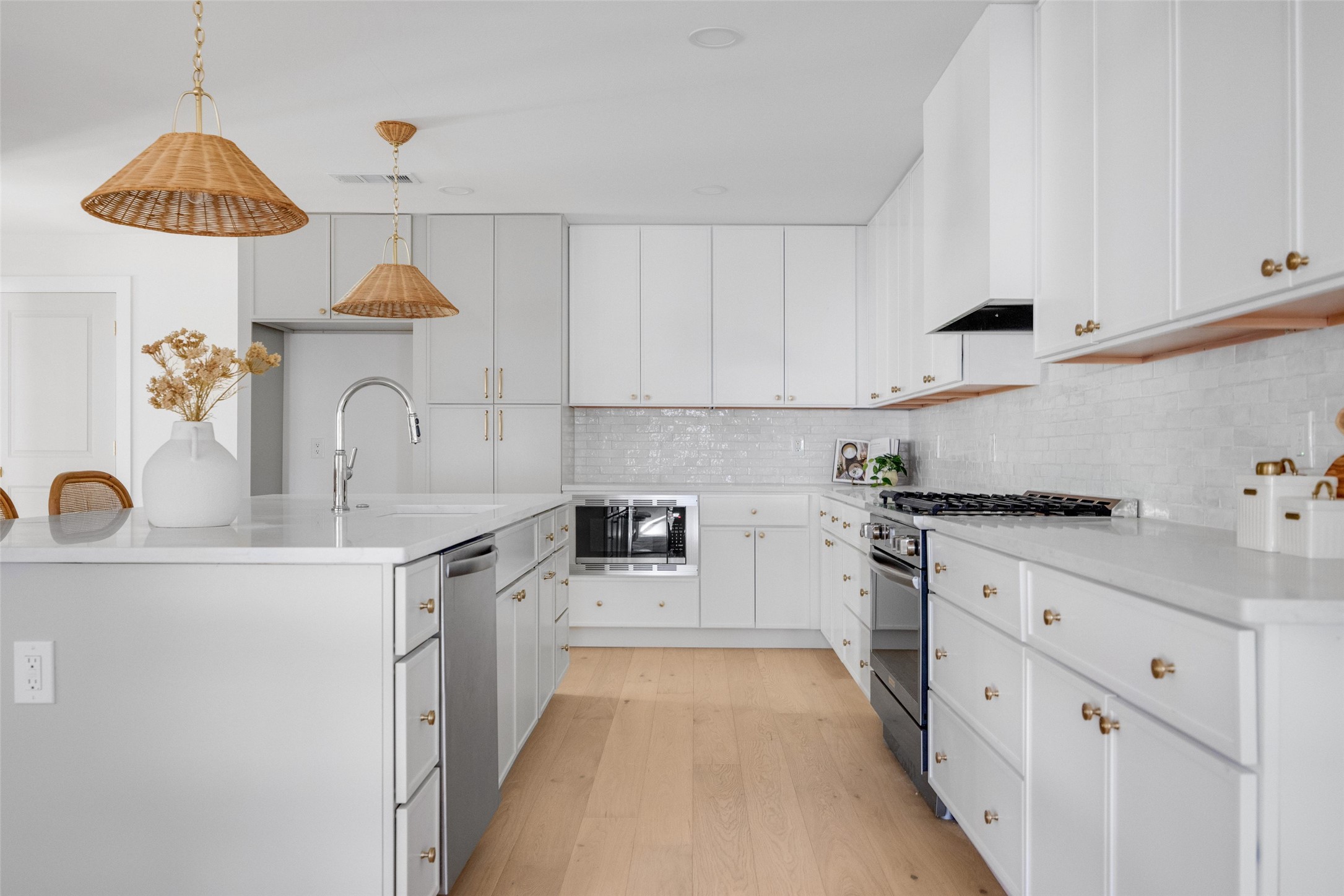 Bright white kitchen with a quartz island, brass cabinet hardware, woven pendant lights, and light wood floors.
