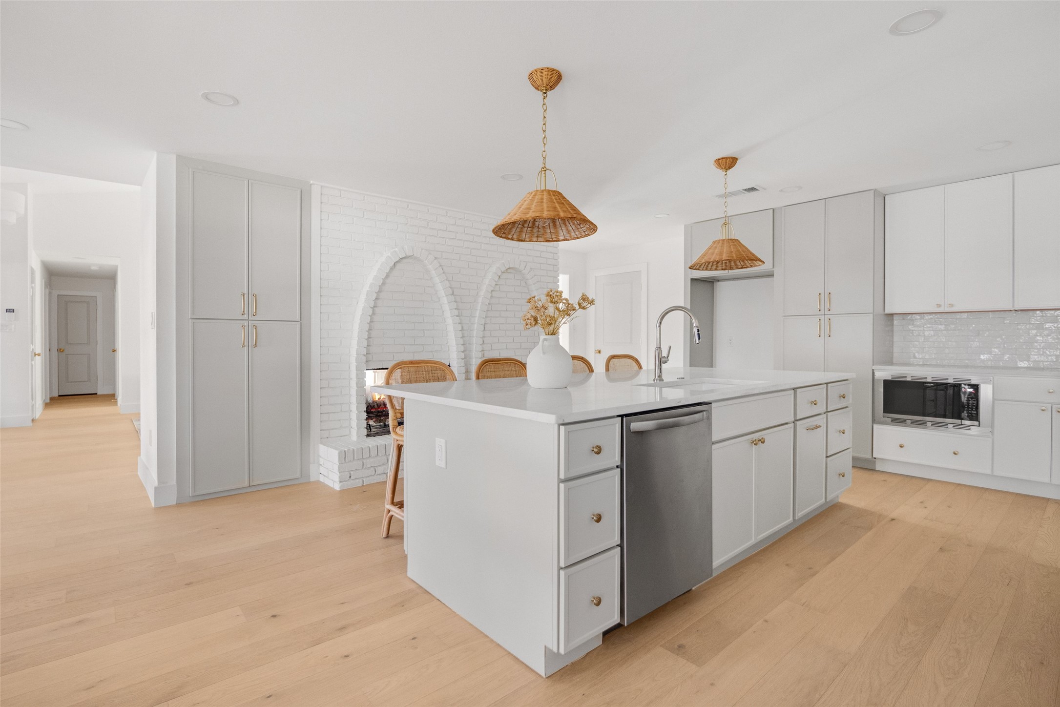 Bright white kitchen with a large island, woven pendant lights, and an arched white brick fireplace.