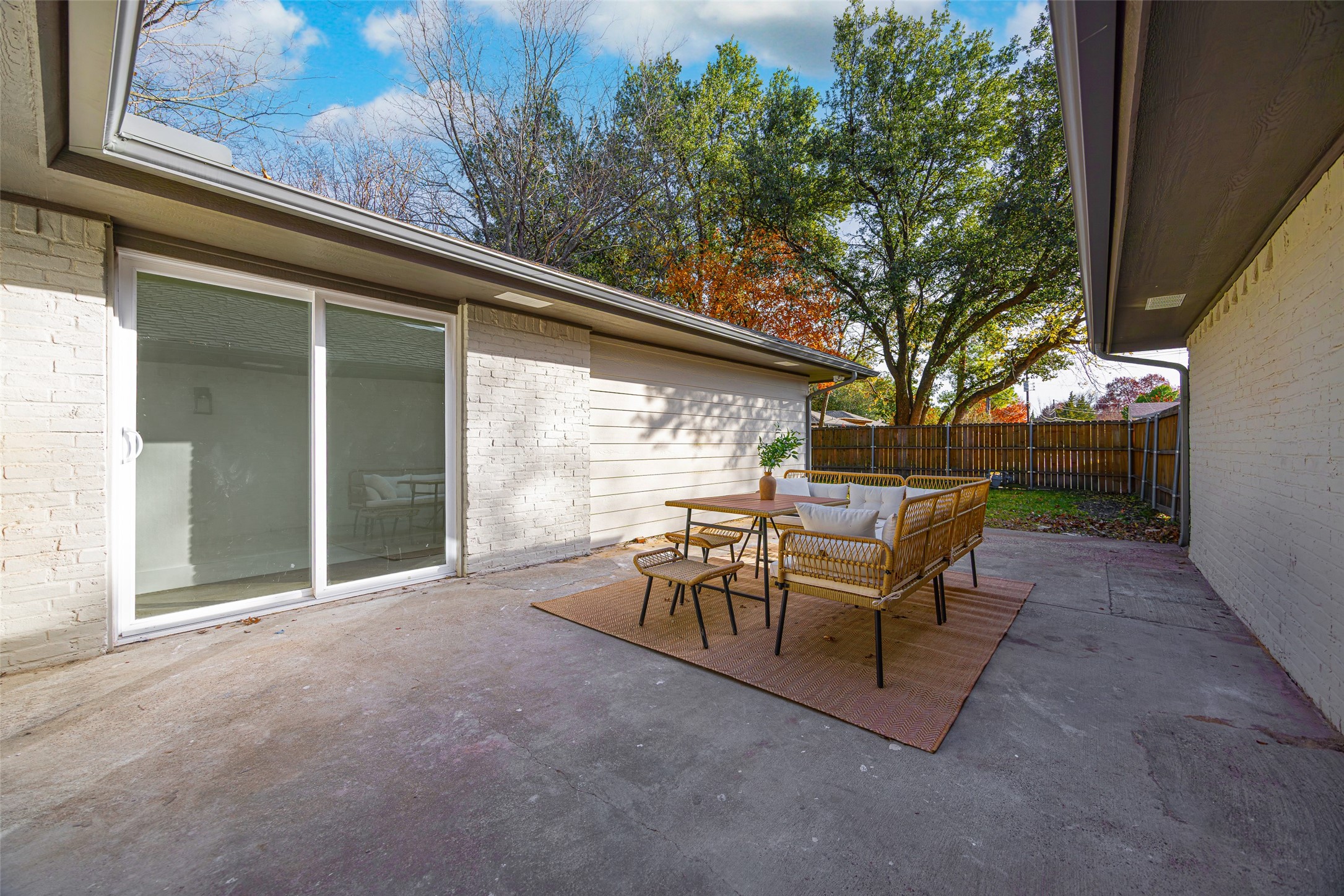 Covered patio with wicker dining and lounge furniture beside a sliding glass door and fenced, tree-shaded yard.