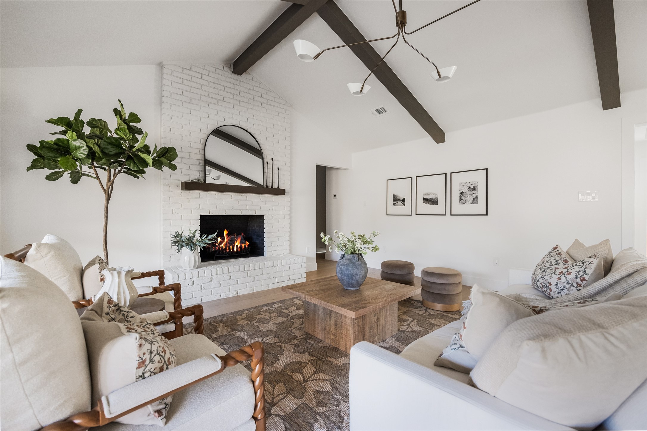 Living room with a white brick fireplace, lit fire, vaulted beamed ceiling, and neutral seating.