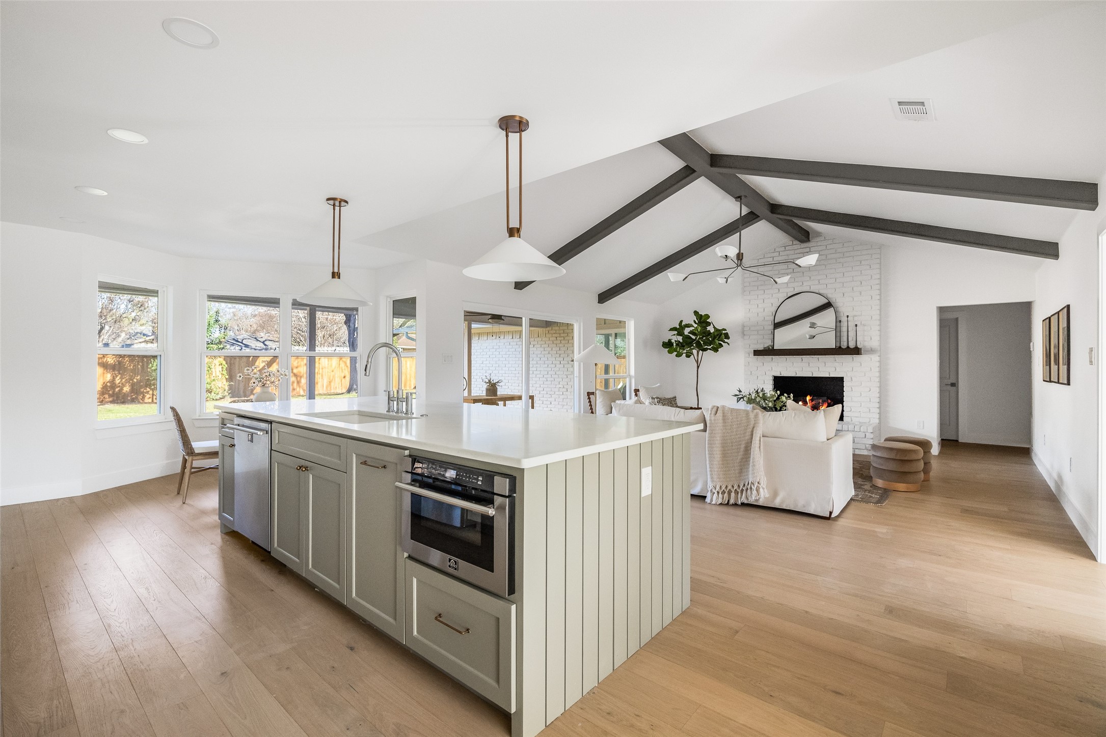 Open-plan kitchen and living room with a large island, vaulted beamed ceiling, and white brick fireplace.