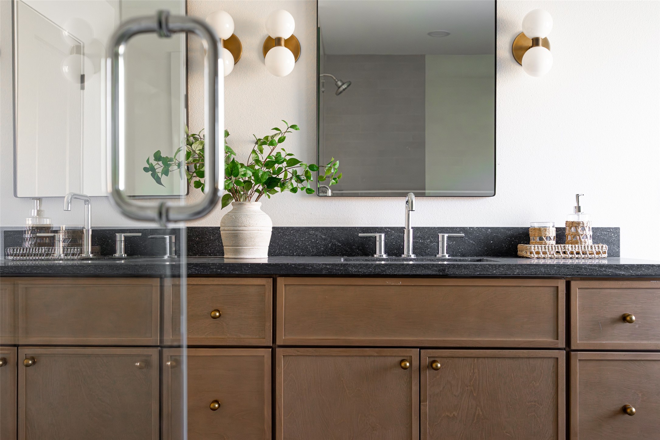 Bathroom vanity with dark stone counters, double sinks, warm wood cabinets, and brass globe sconces.