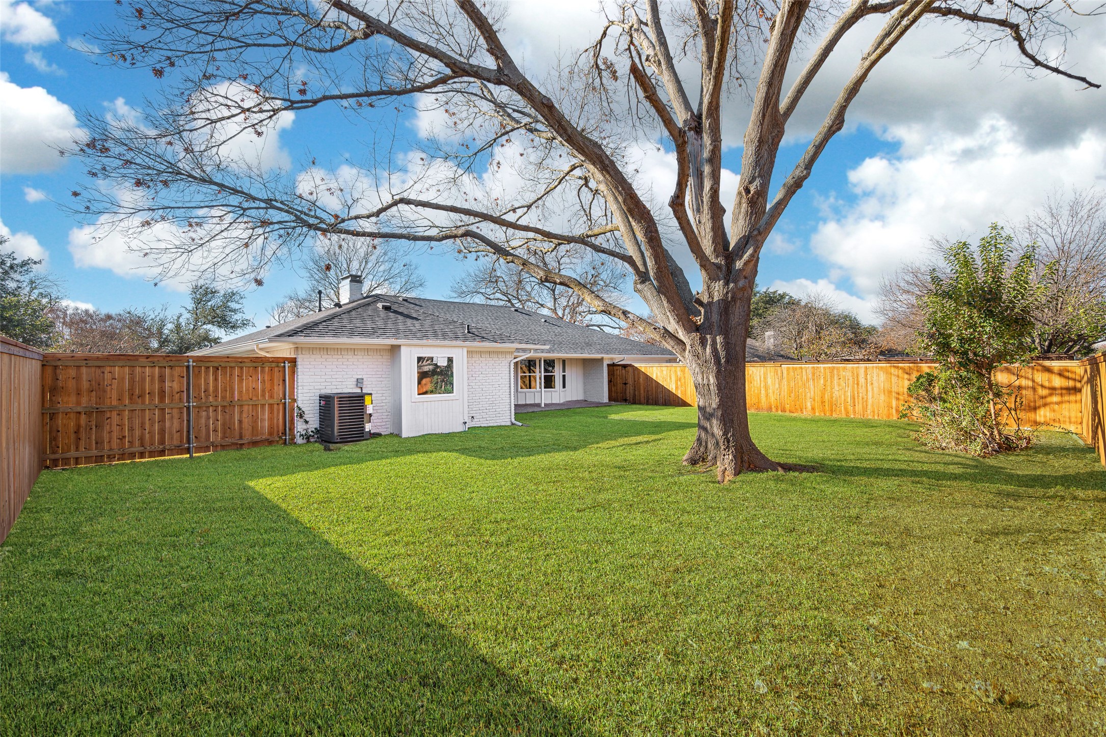 White brick home behind a broad lawn, with a large bare tree and wooden privacy fence.