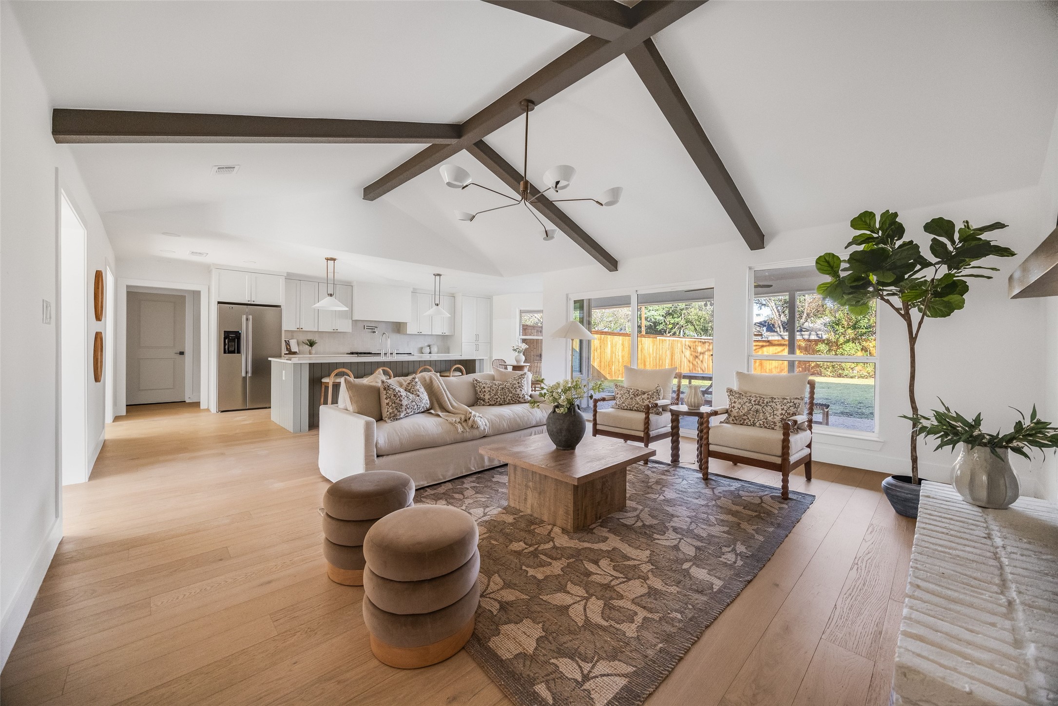 Open-plan living room and kitchen with vaulted beamed ceiling, neutral furnishings, and large backyard-facing windows.