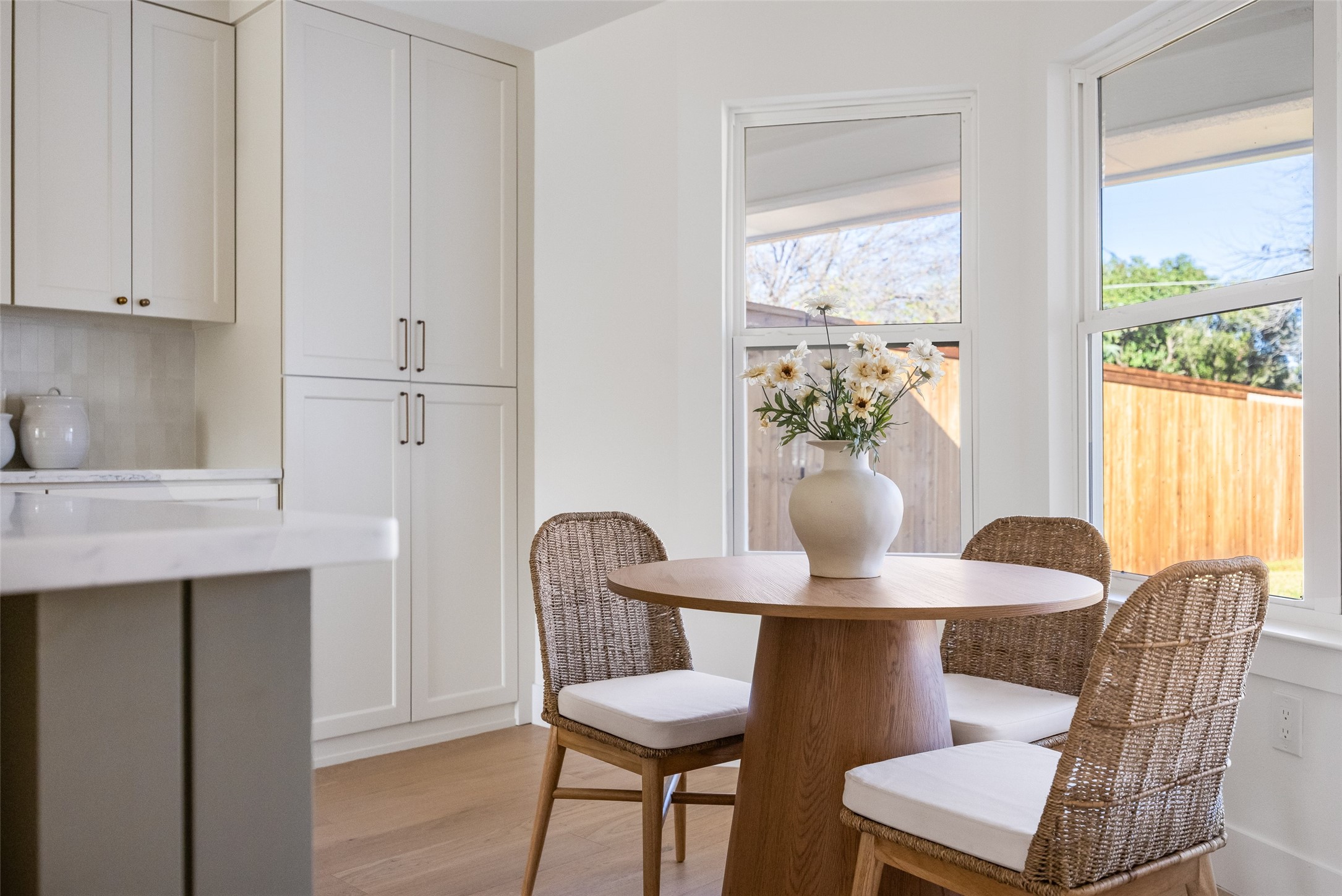 Kitchen dining nook with a round wood table, wicker chairs, and large windows overlooking a fenced yard.