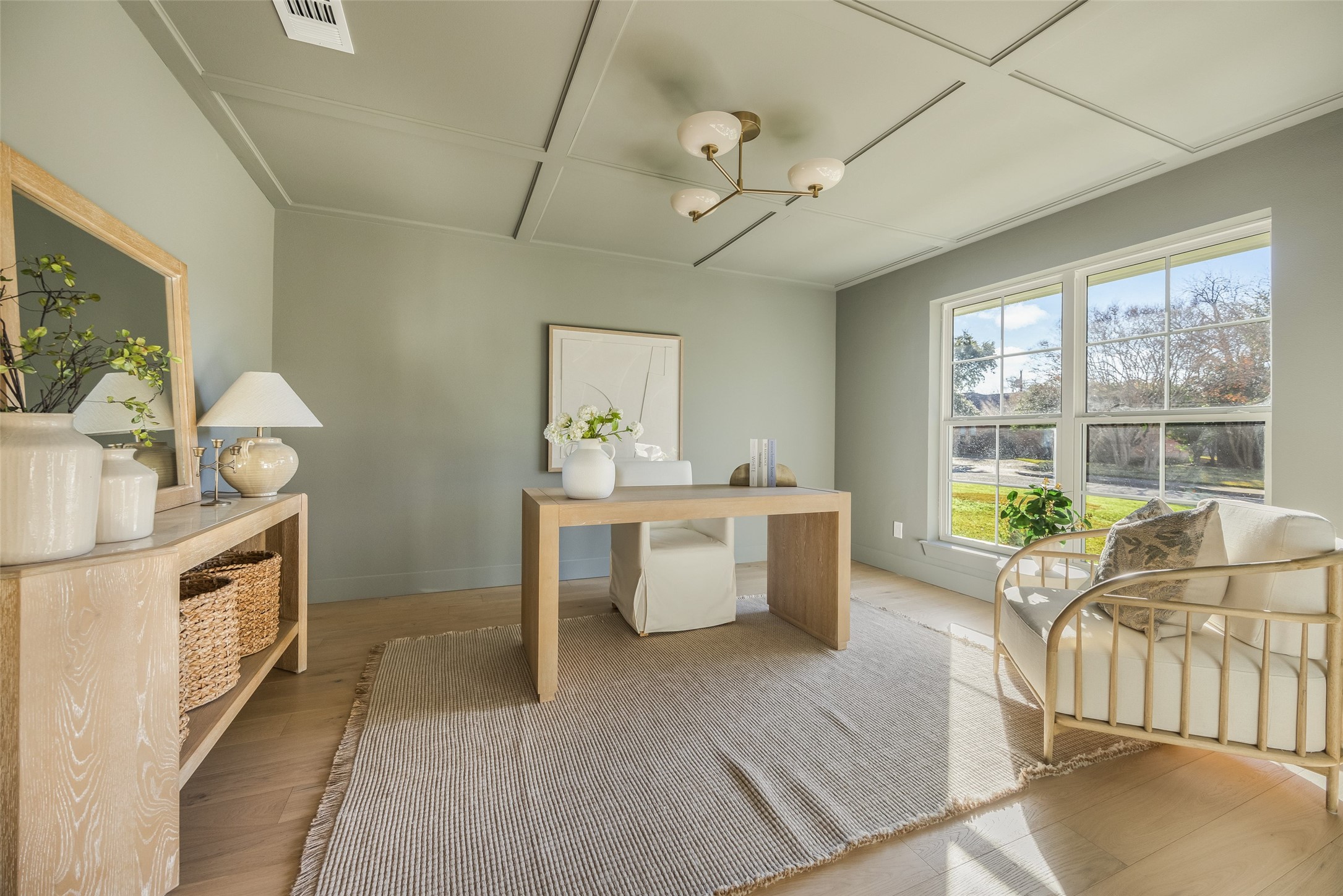 Staged home office with a light wood desk, textured rug, and armchair beside large windows.