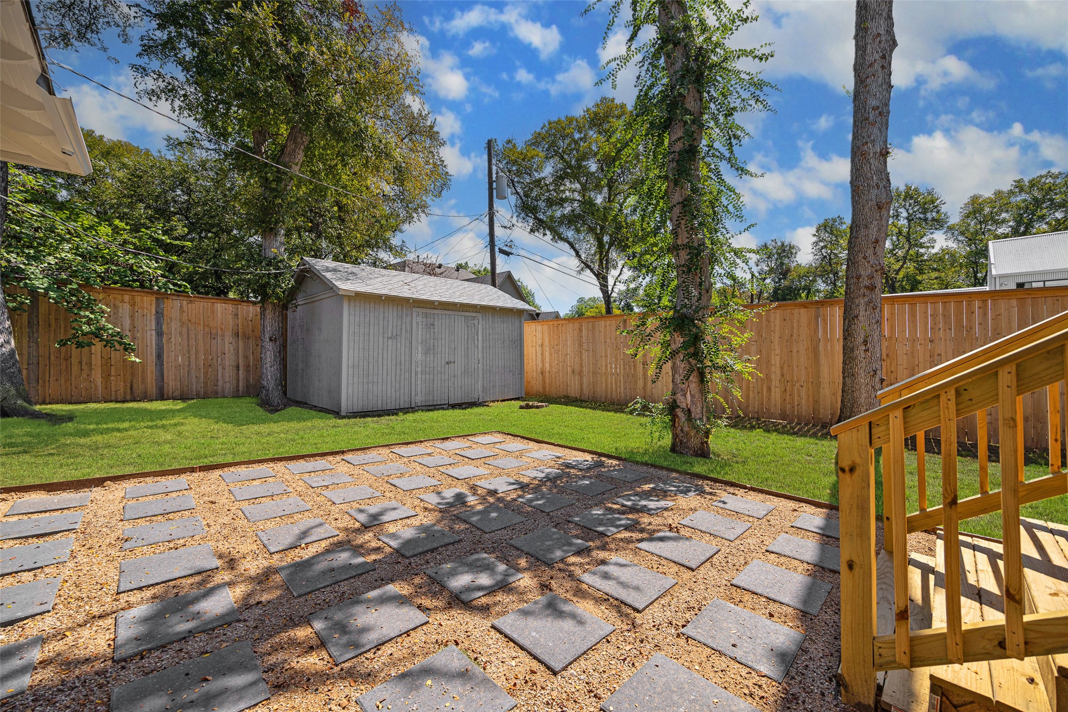 Backyard patio with square stepping stones, a gray shed, mature trees, and wooden deck steps.