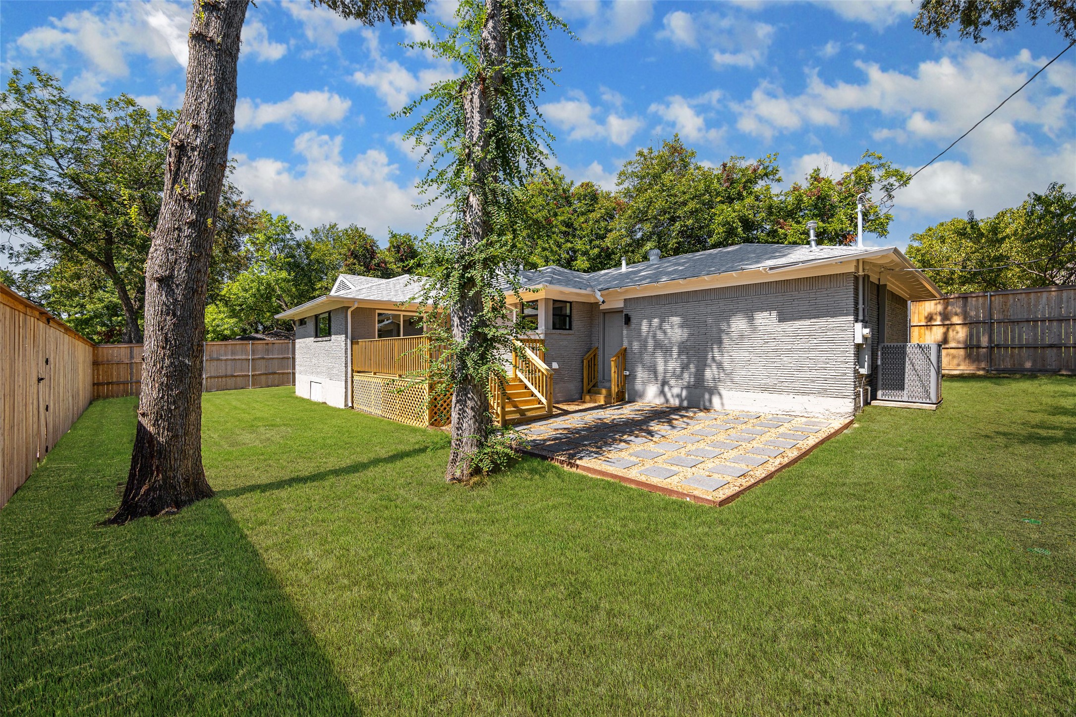 Rear of a gray brick house with a raised wooden deck, paved patio, and fenced grassy yard.