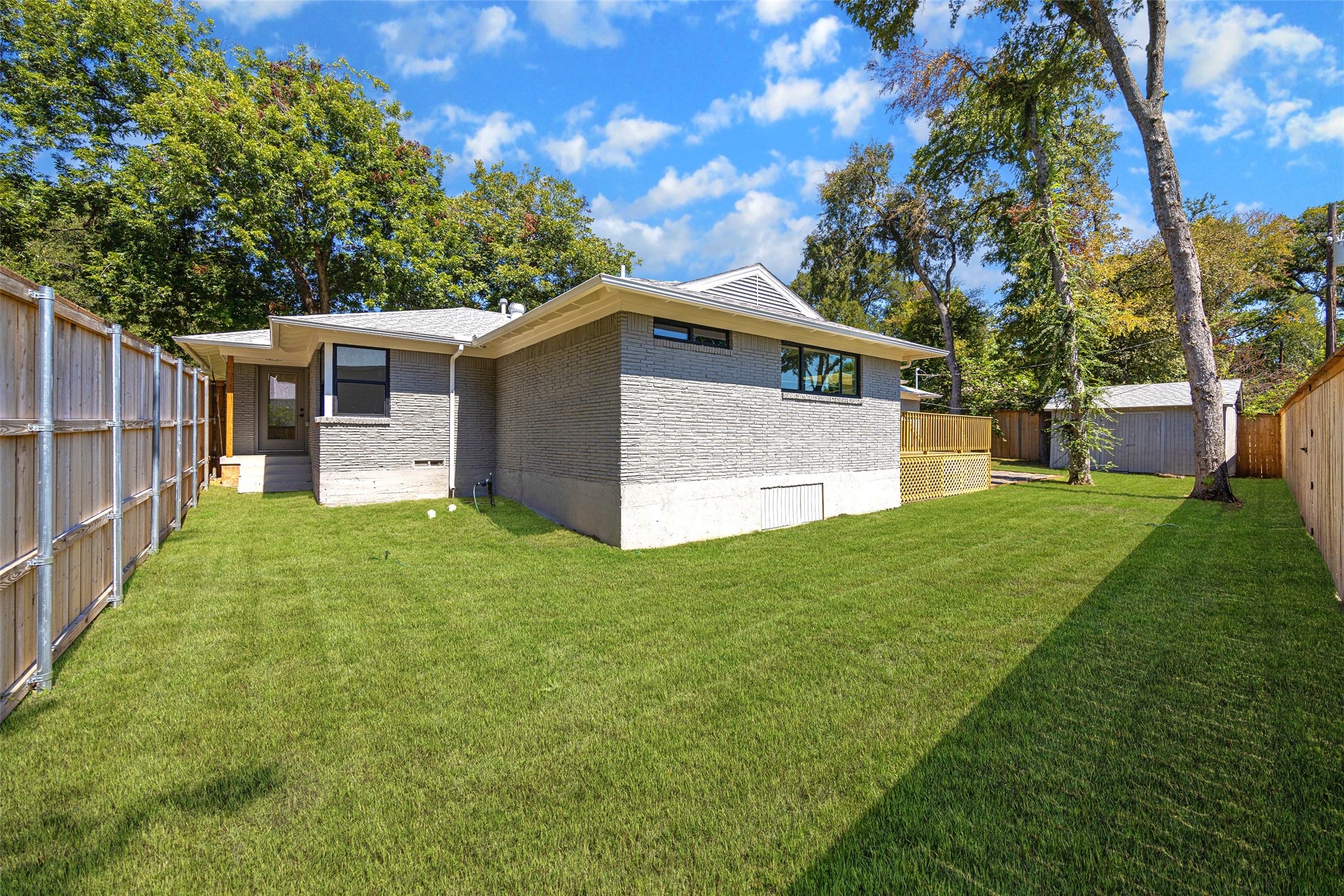 Gray brick home with a spacious fenced lawn, mature trees, and a detached shed.