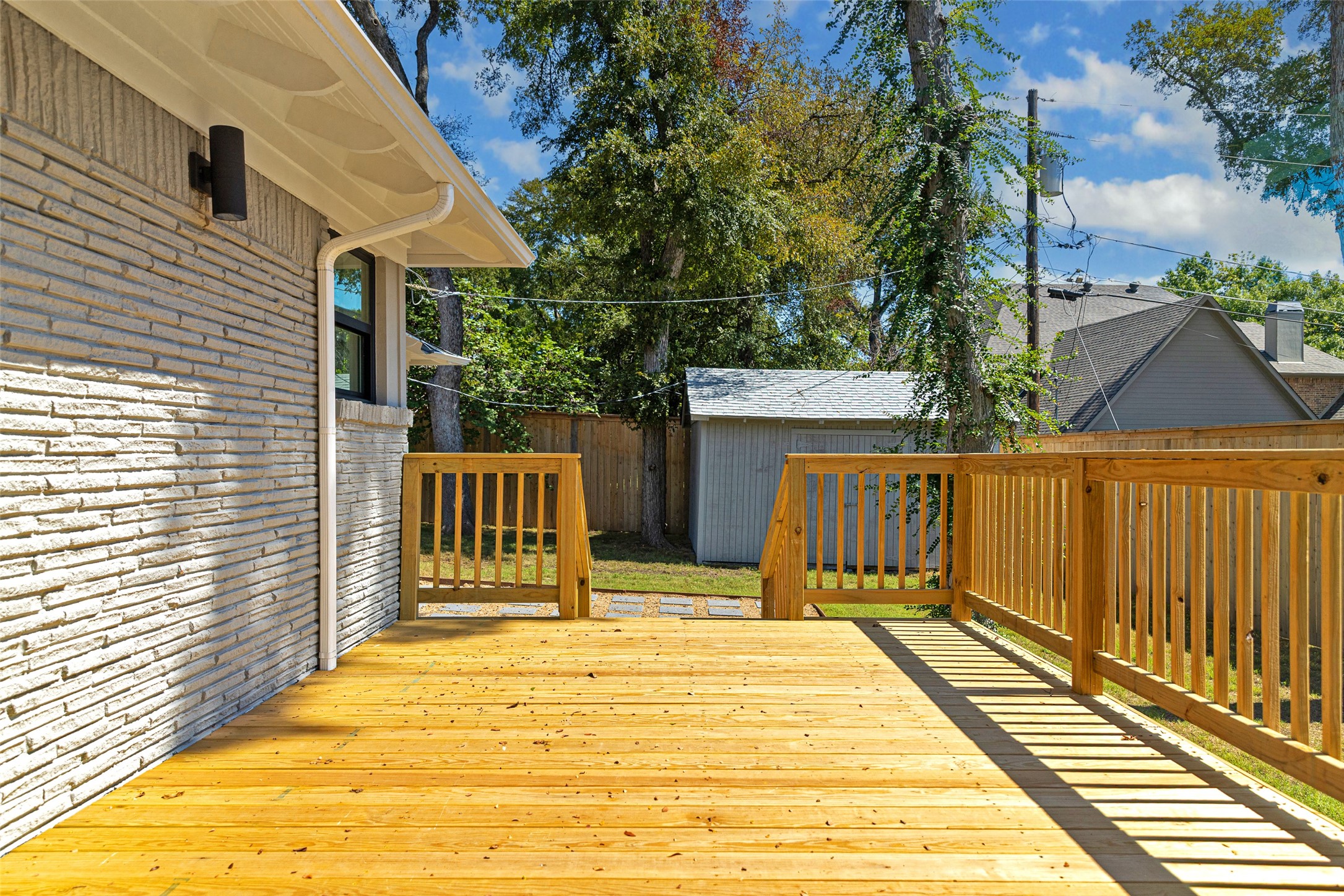 Wood backyard deck with railings beside a brick house, overlooking a lawn, shed, and mature trees.