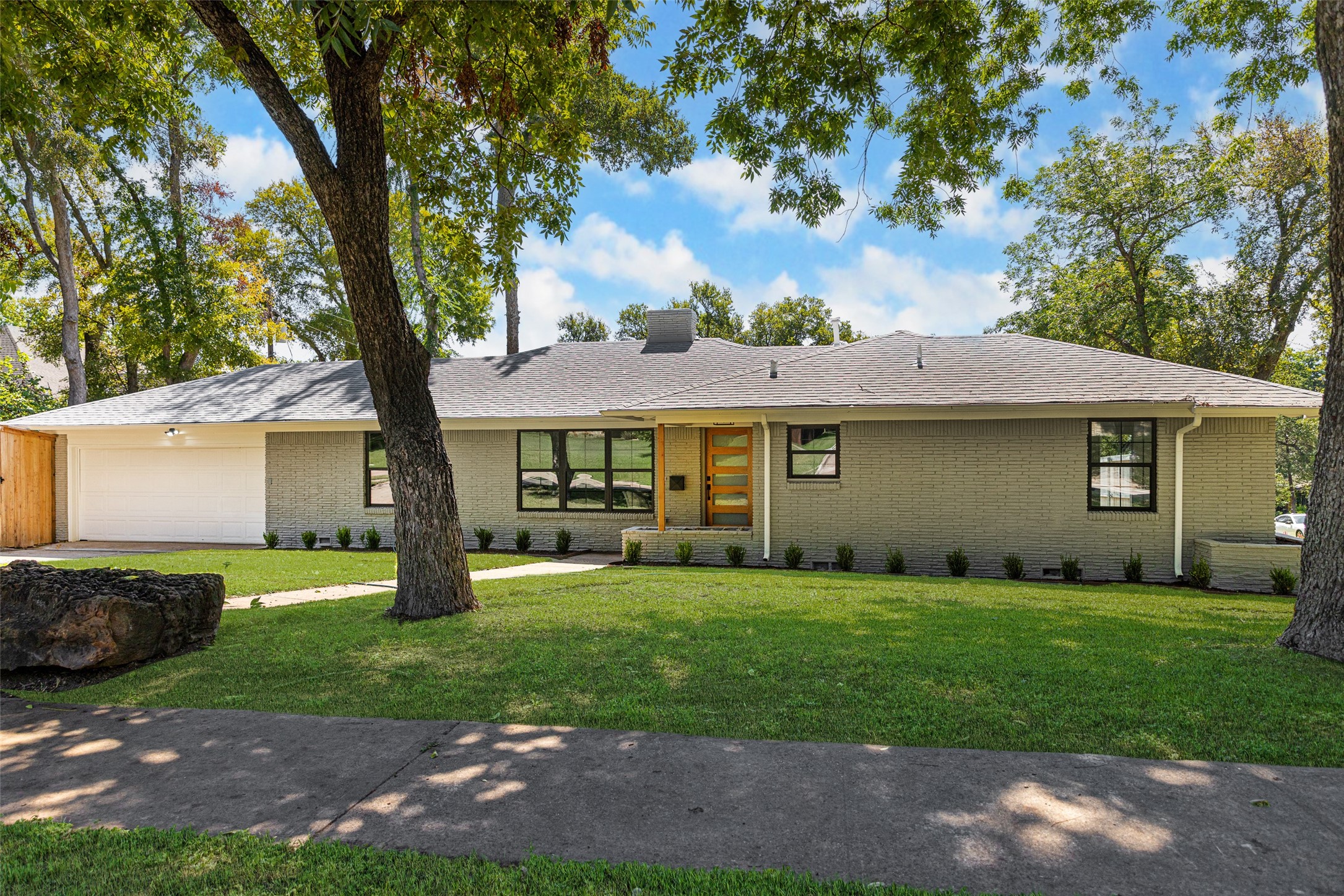 Single-story gray-brick home with an attached garage, modern wood entry door, and mature trees across the lawn.