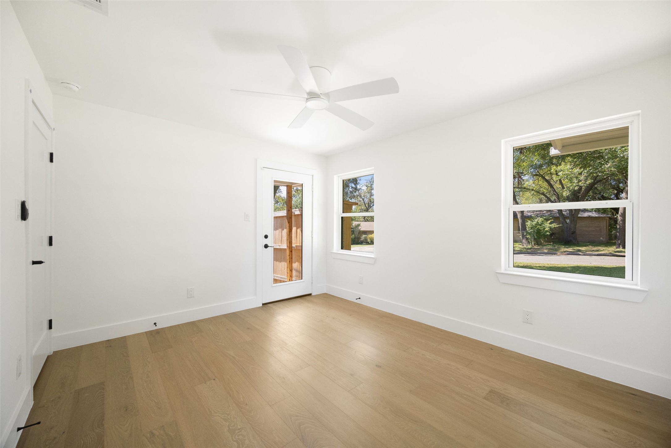 Vacant room with light wood floors, a ceiling fan, and windows overlooking leafy trees.