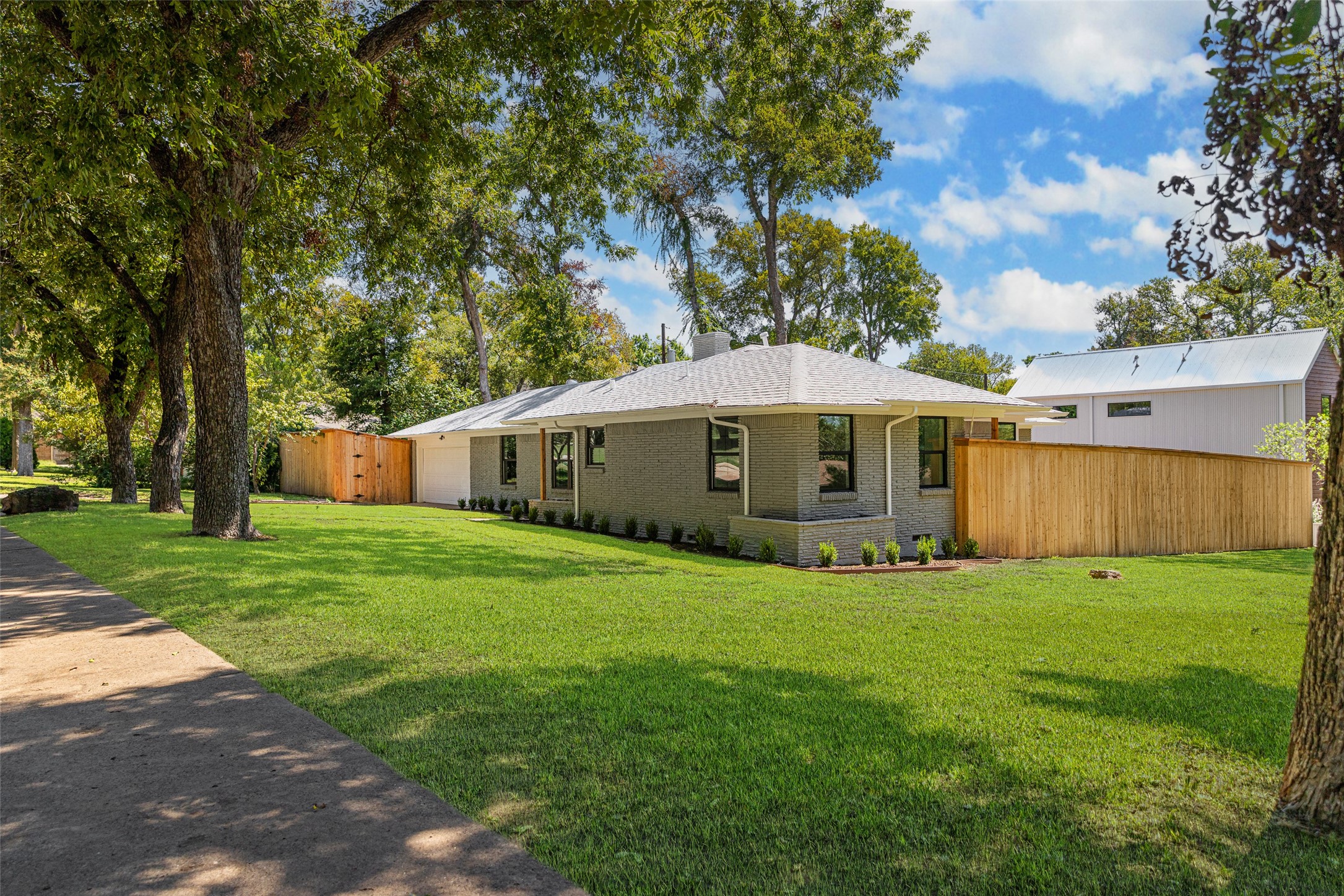 Single-story brick house behind a broad lawn, framed by mature trees and a wooden privacy fence.