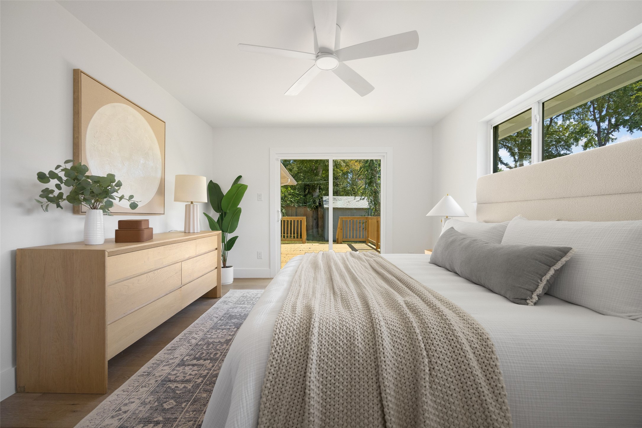Staged bedroom with a white bed and textured blanket, light wood dresser, and sliding doors to a deck.