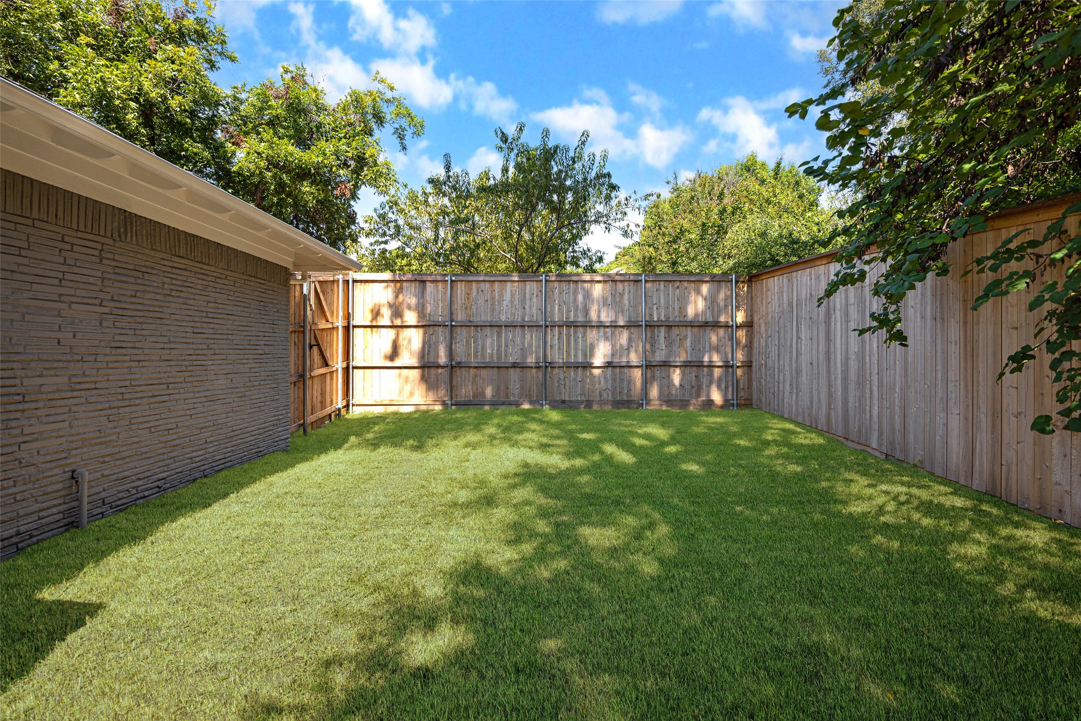 Fenced backyard with a broad green lawn beside a brick house and mature trees.