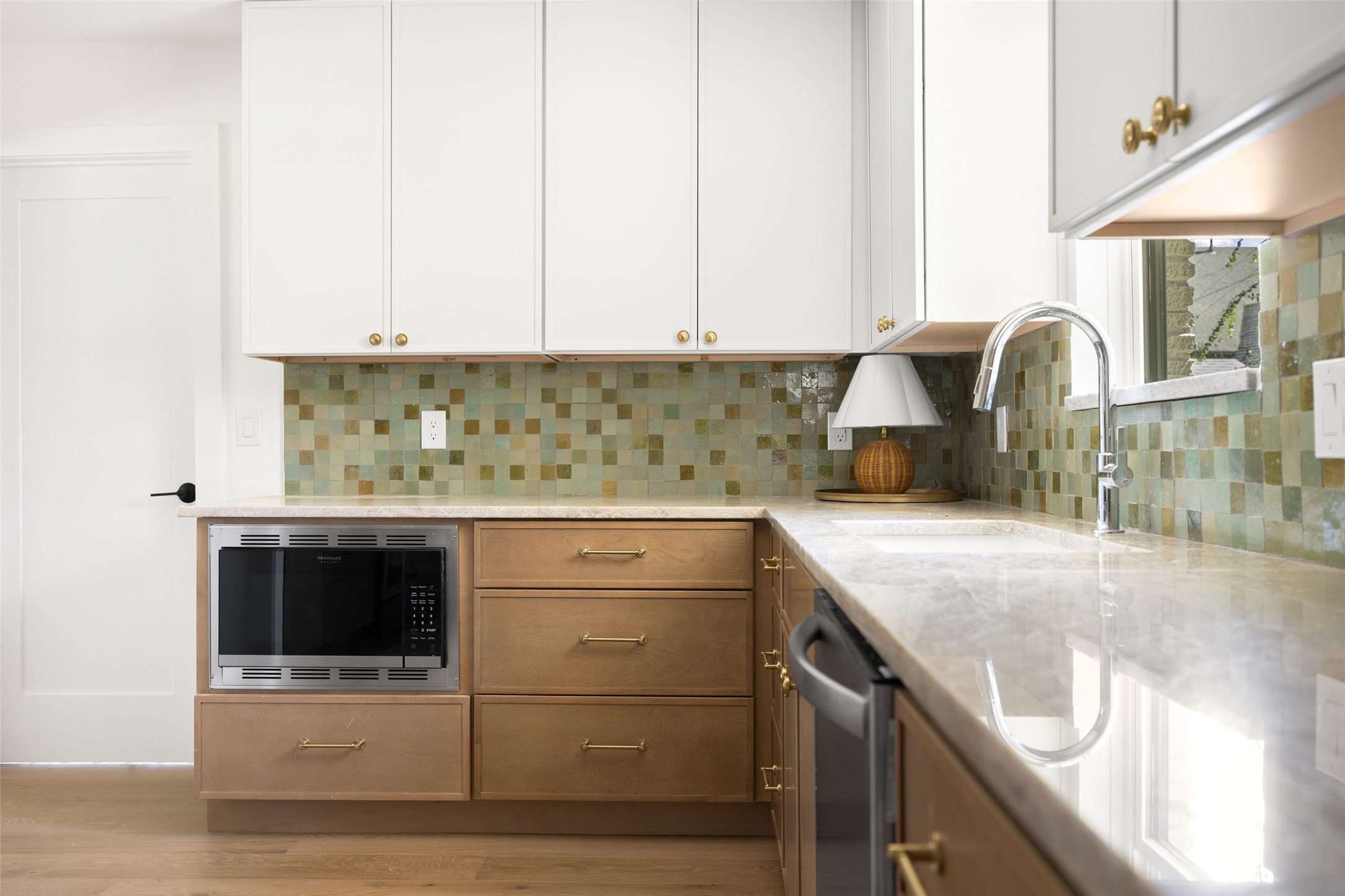 Kitchen with green mosaic backsplash, warm wood cabinets, and a large chrome faucet beside a window.