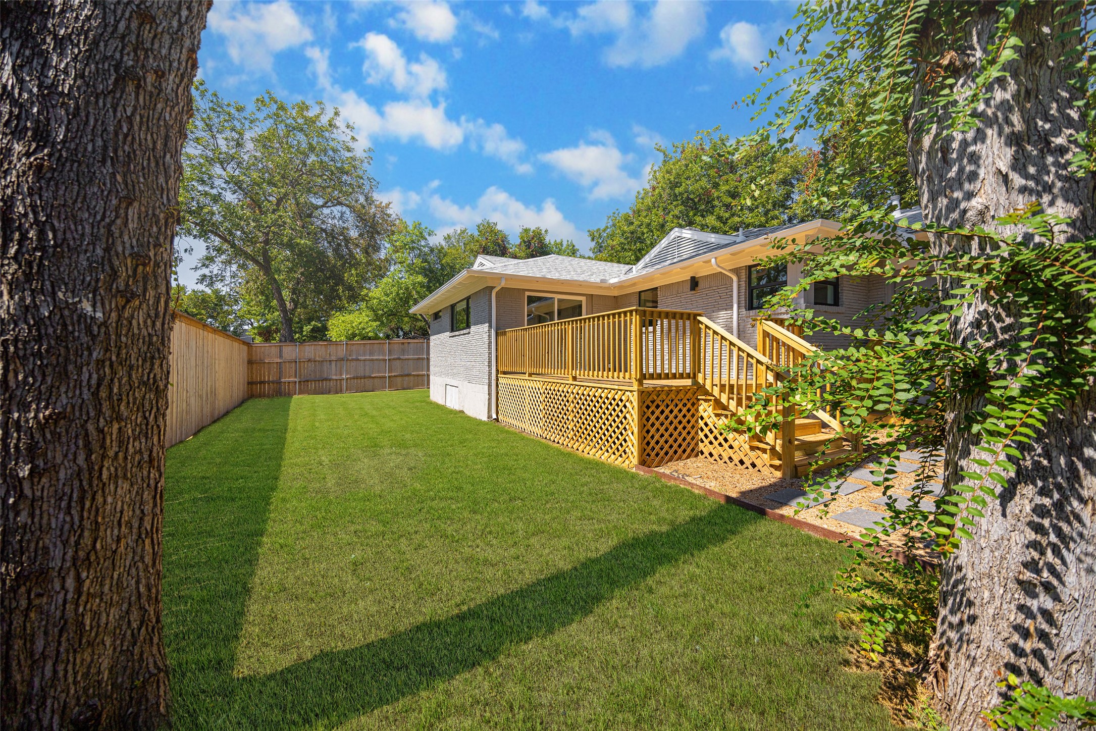 Fenced backyard with a wide lawn and raised wooden deck framed by mature trees.