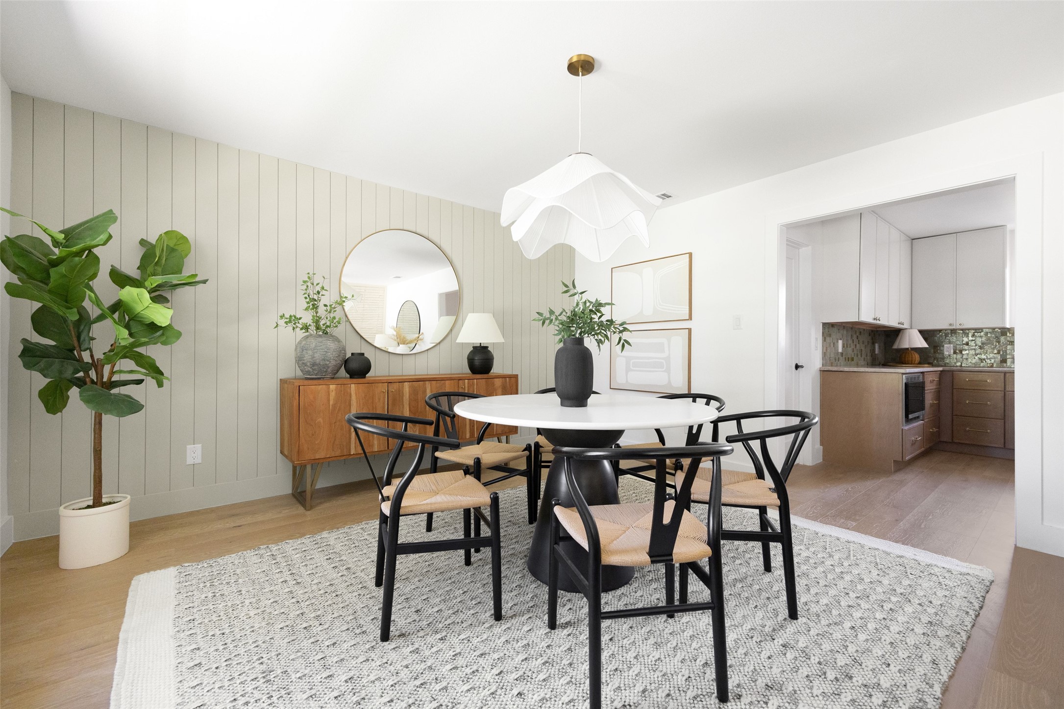 Dining room with a round white table, black chairs, paneled wall, and open kitchen.