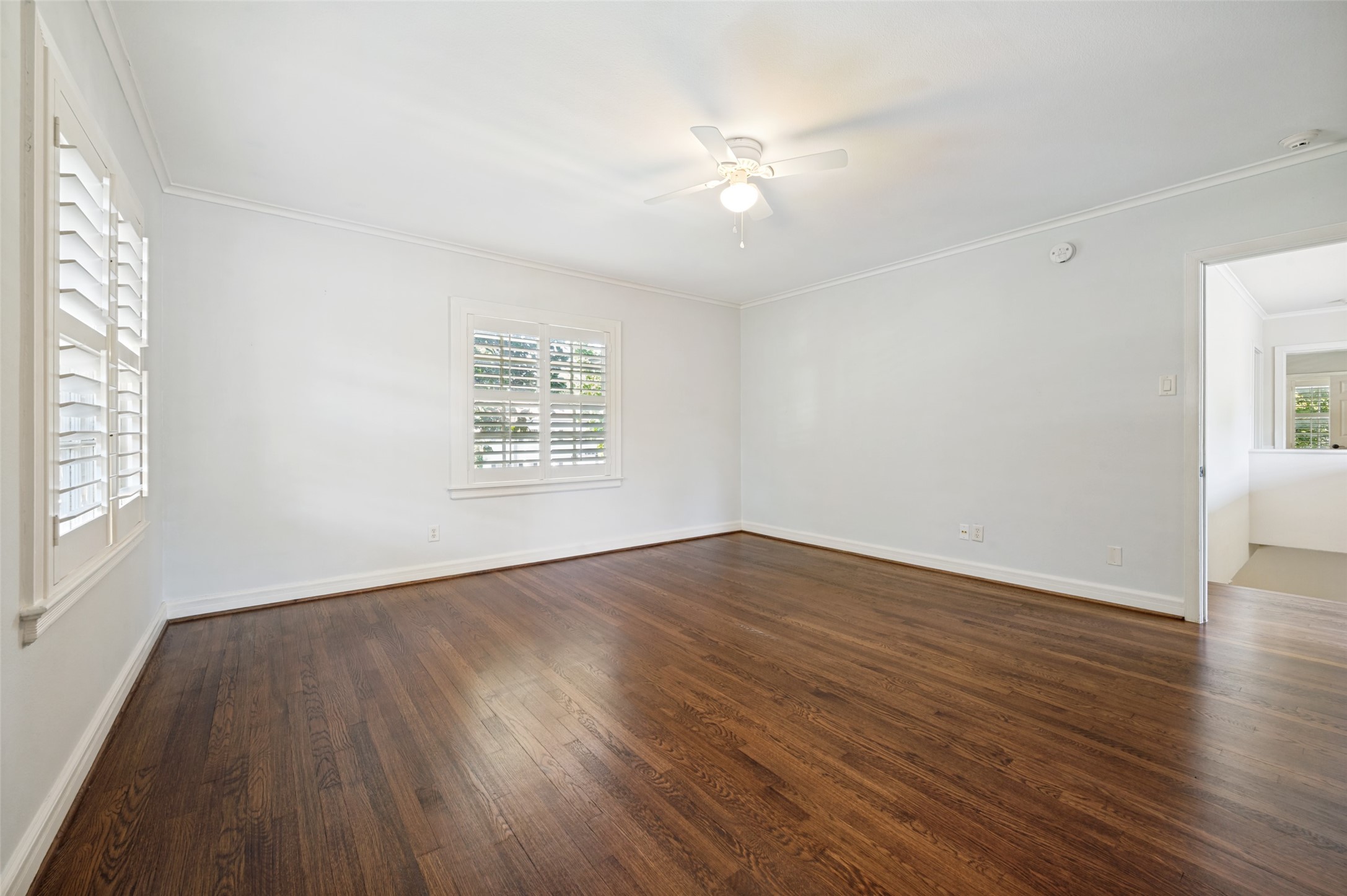 Vacant room with dark hardwood floors, plantation shutters, and a ceiling fan.