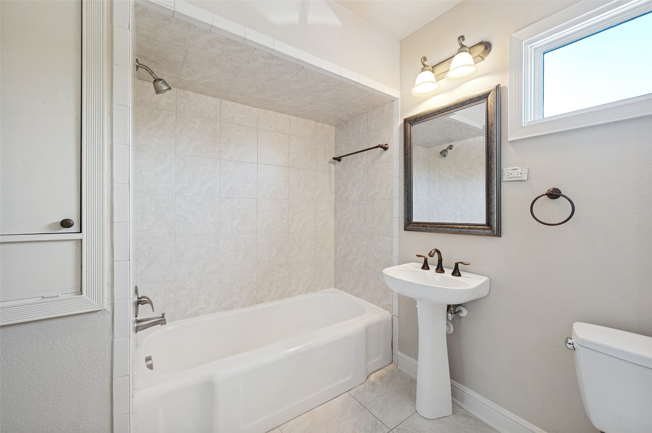 Bathroom with a white bathtub, tiled shower surround, and pedestal sink beside a window.