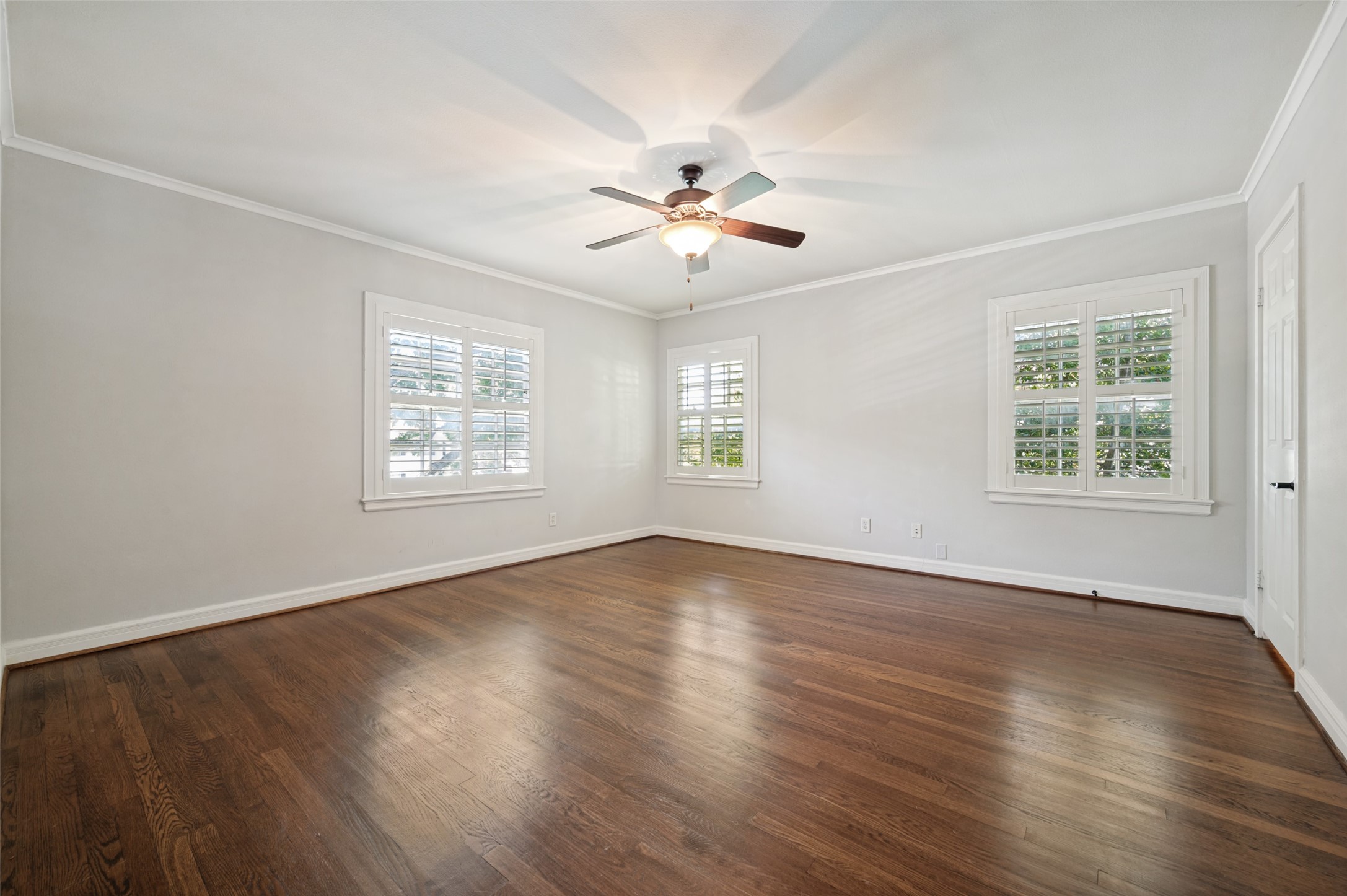 Empty room with dark hardwood floors, three shuttered windows, and a ceiling fan.