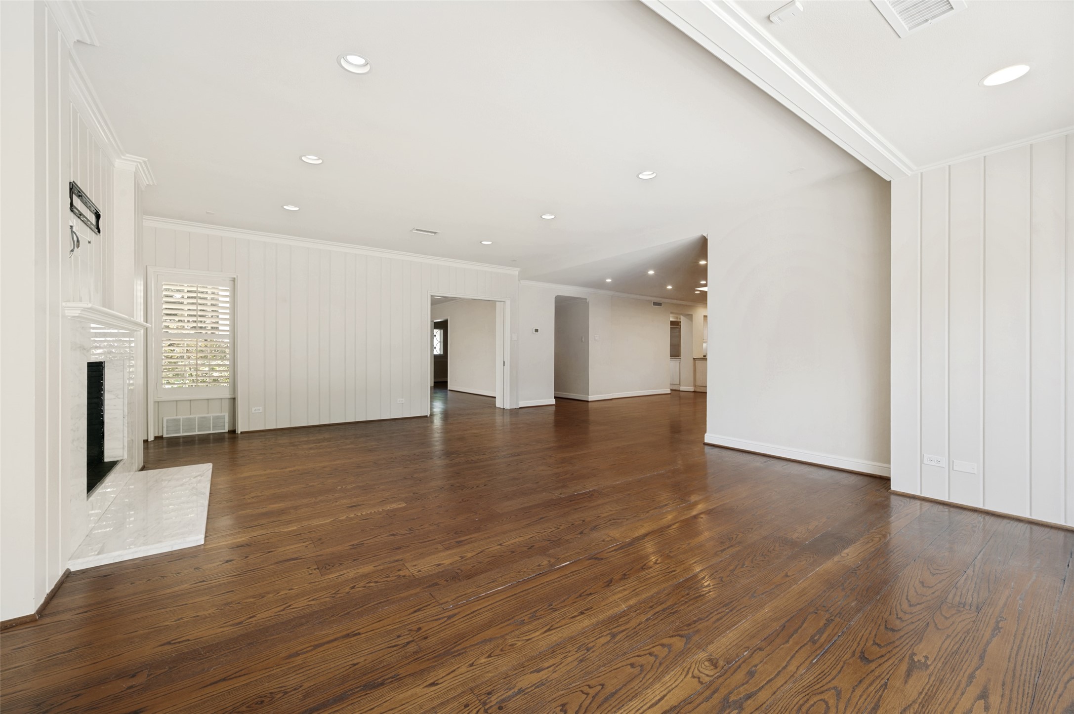 Empty living room with hardwood floors, a marble fireplace, and plantation shutters.