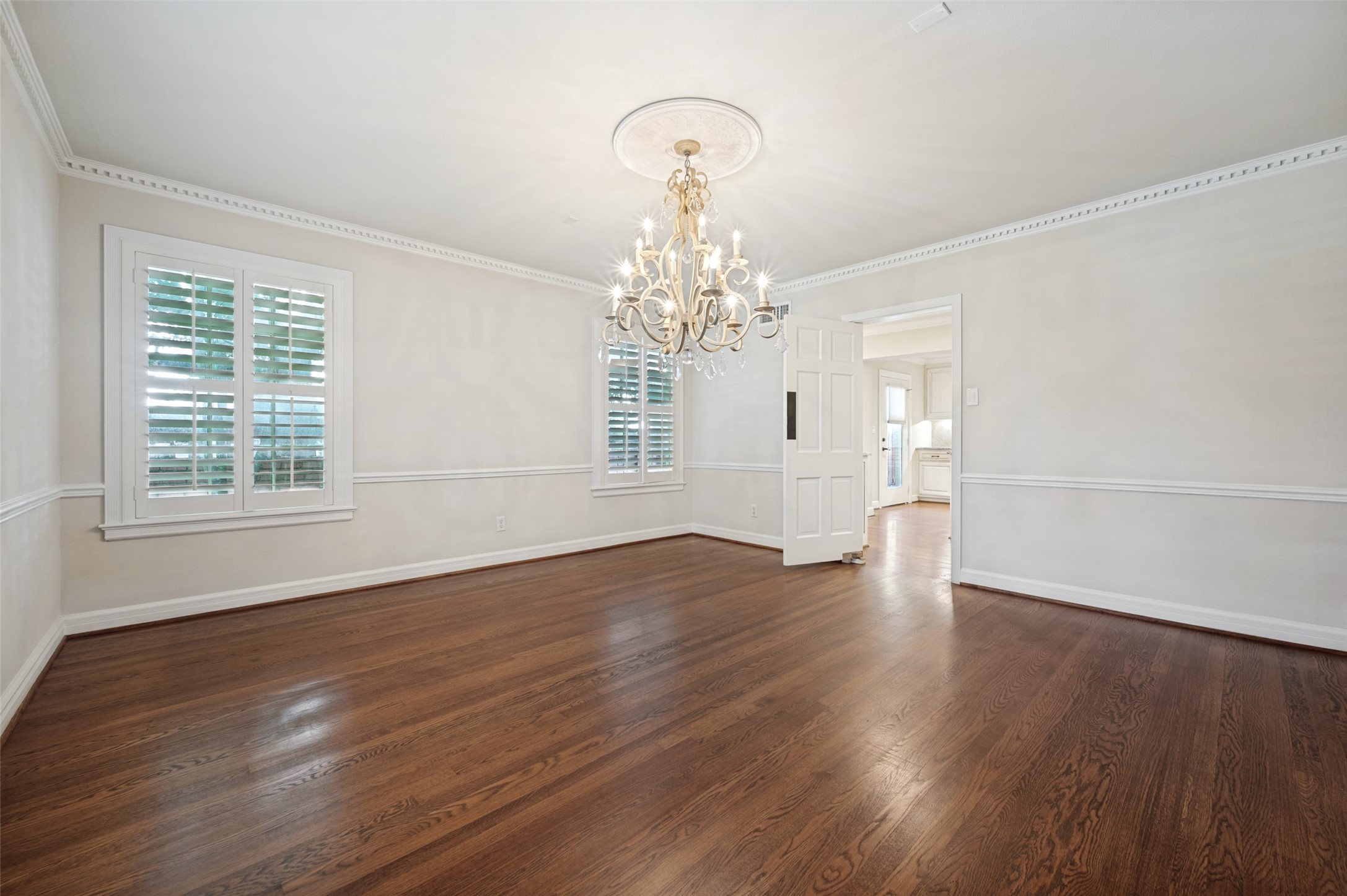 Empty dining room with dark hardwood floors, plantation shutters, and an ornate chandelier.