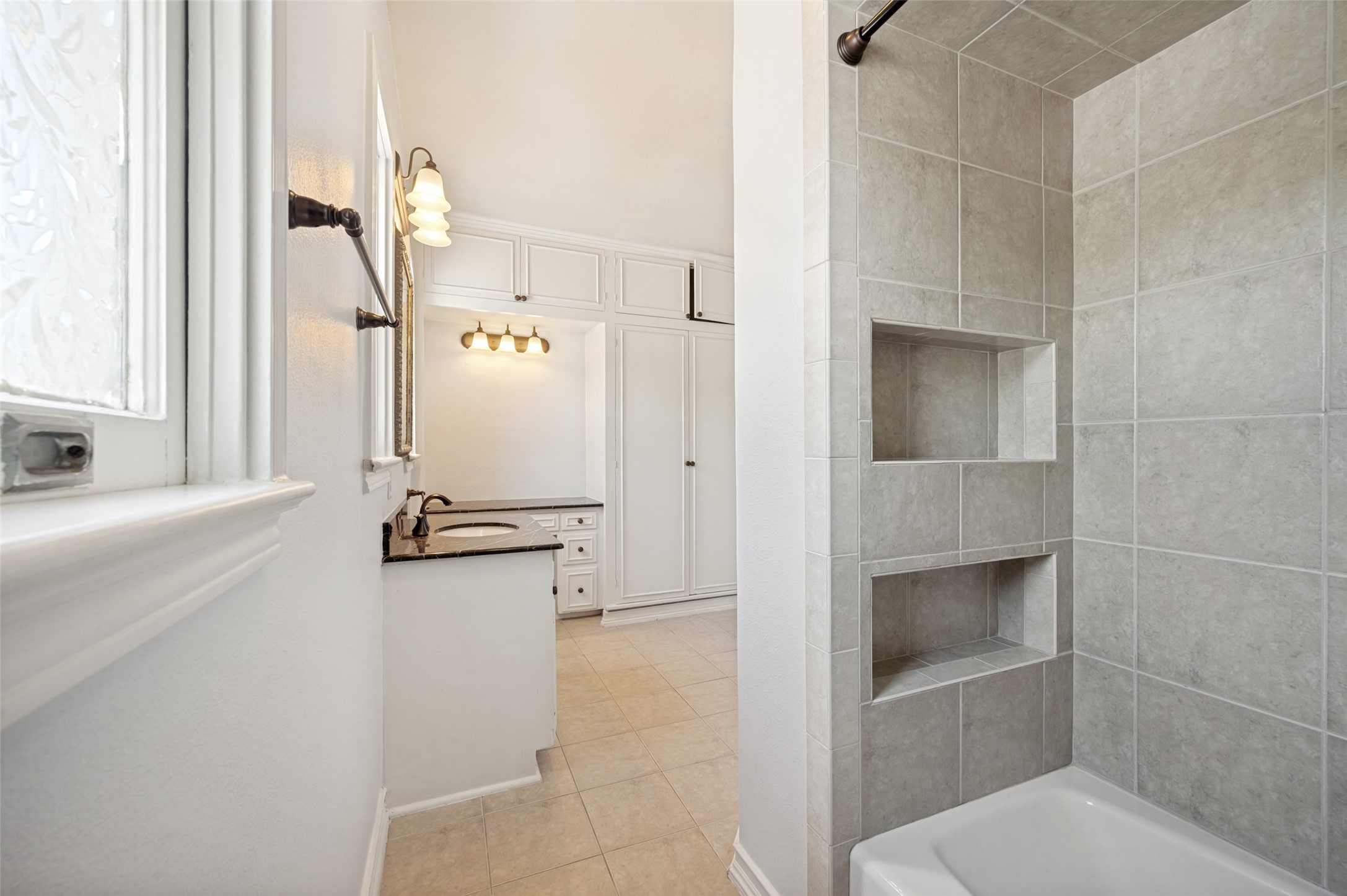 Bathroom with gray tiled shower niches beside a white vanity and built-in cabinets.