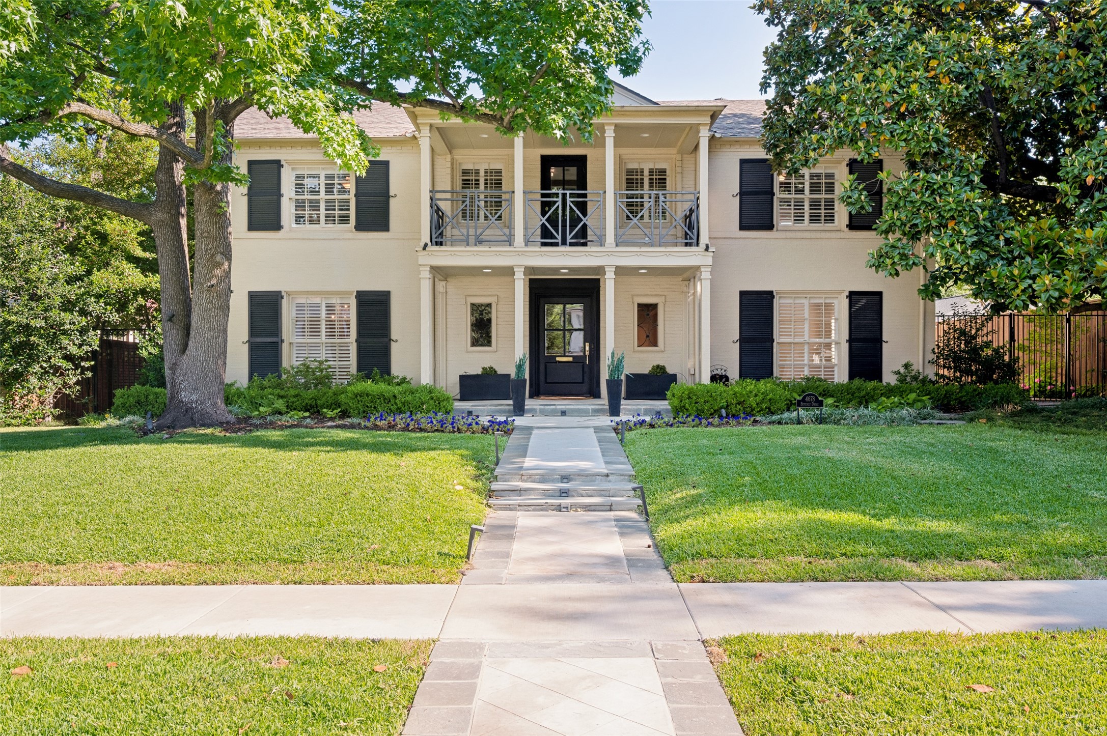 Two-story cream house with black shutters, a balcony, mature trees, and a stone front walkway.