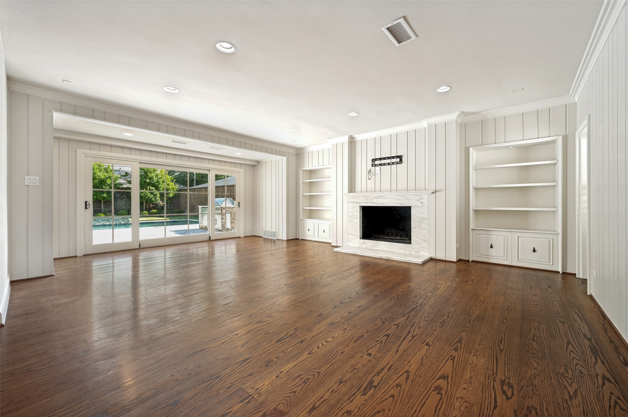 Empty living room with hardwood floors, marble fireplace, built-in shelves, and sliding doors overlooking a pool.