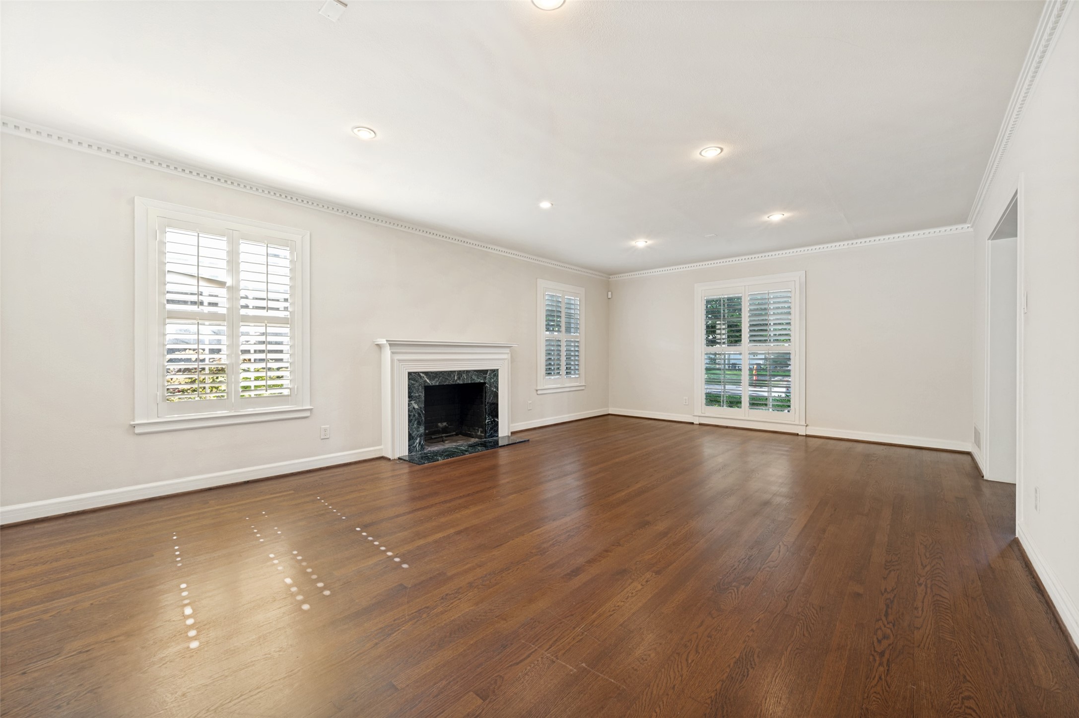 Empty living room with hardwood floors, marble fireplace, and plantation shutters.