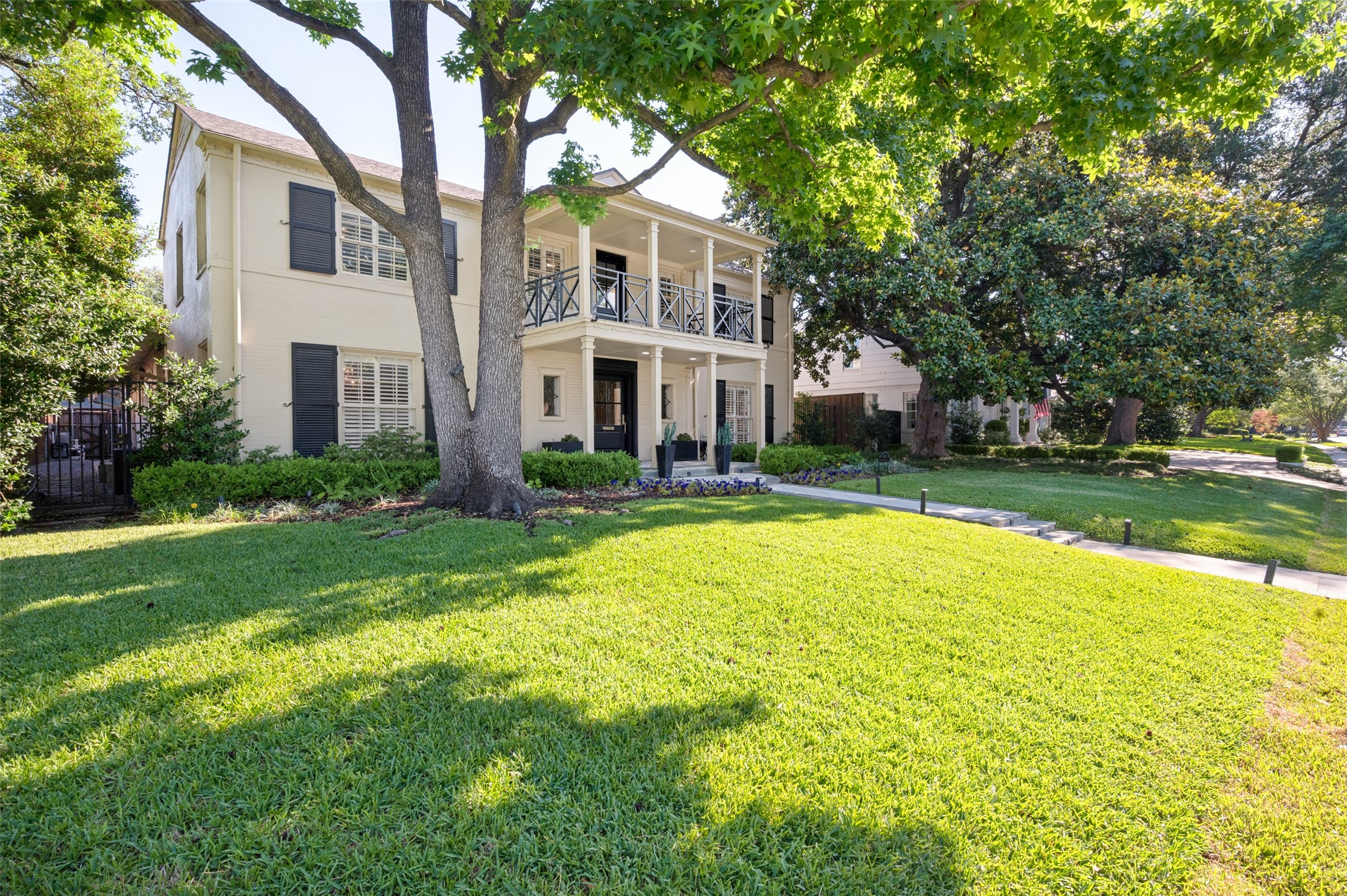 Two-story cream house with an upper balcony, framed by mature trees and a broad green lawn.