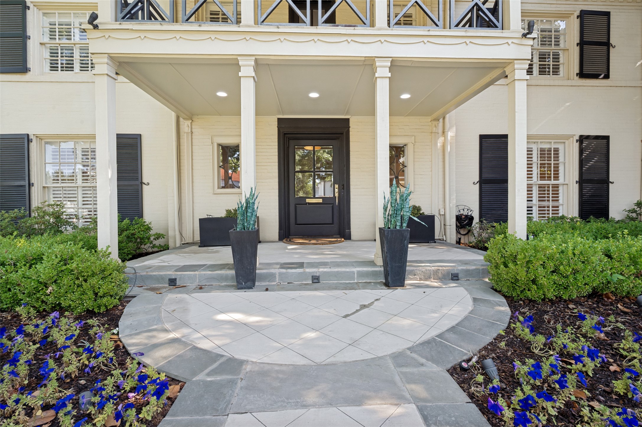 White brick home’s covered front porch with black door, curved stone walkway, and blue flower beds.