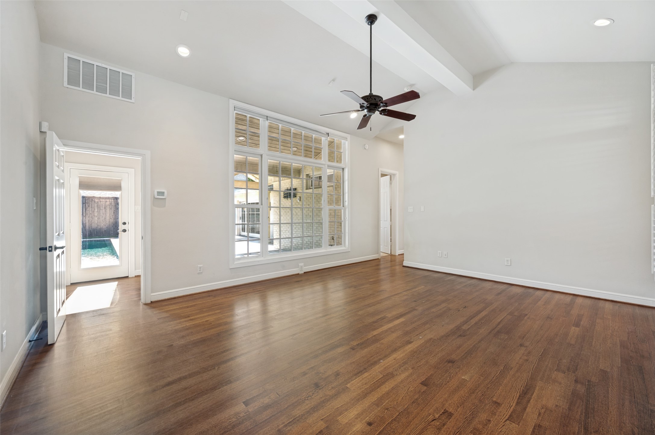 Empty living room with hardwood floors, a ceiling fan, and large windows overlooking a pool.
