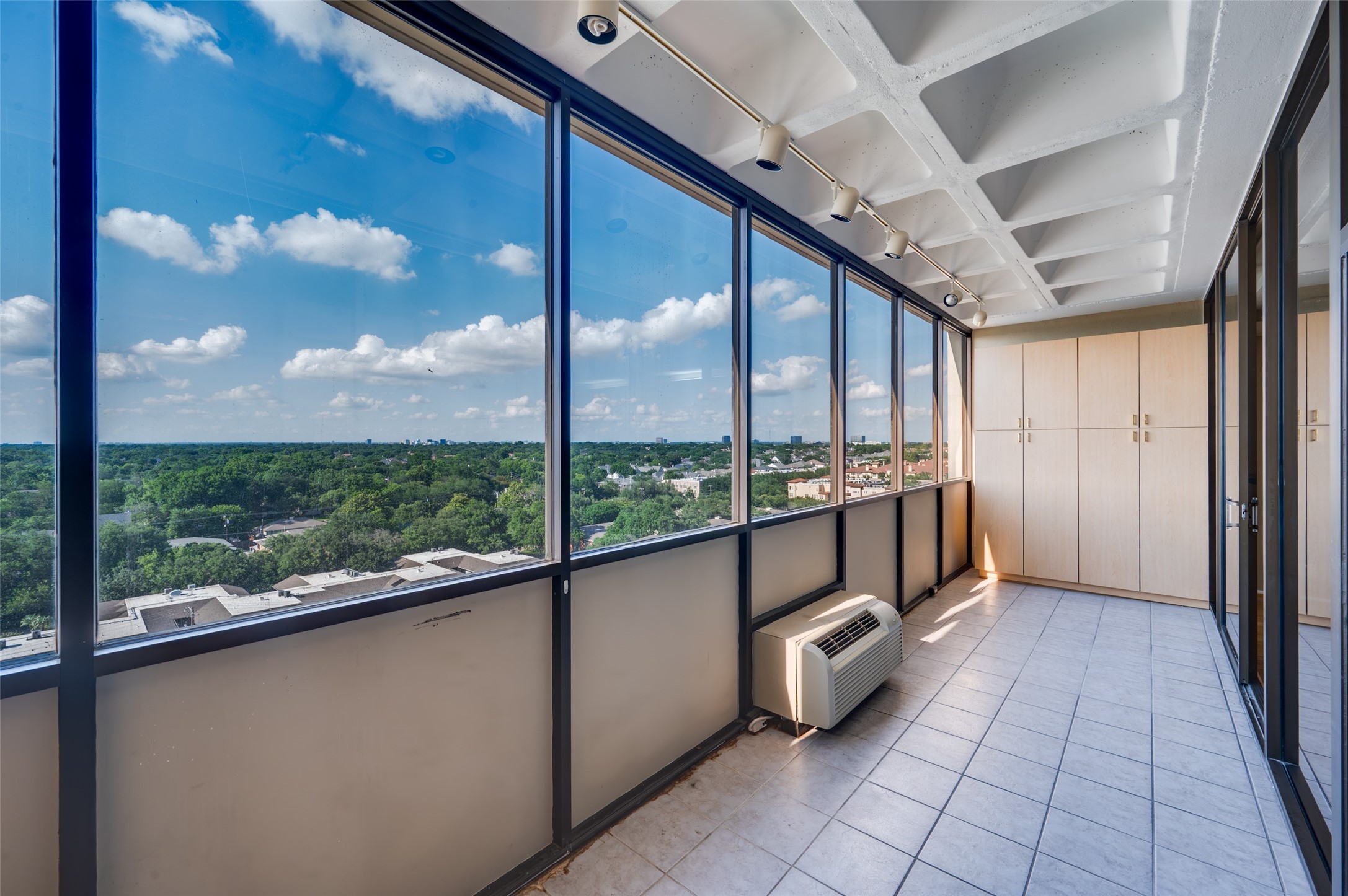 Enclosed sunroom with floor-to-ceiling windows overlooking a leafy neighborhood and distant skyline.