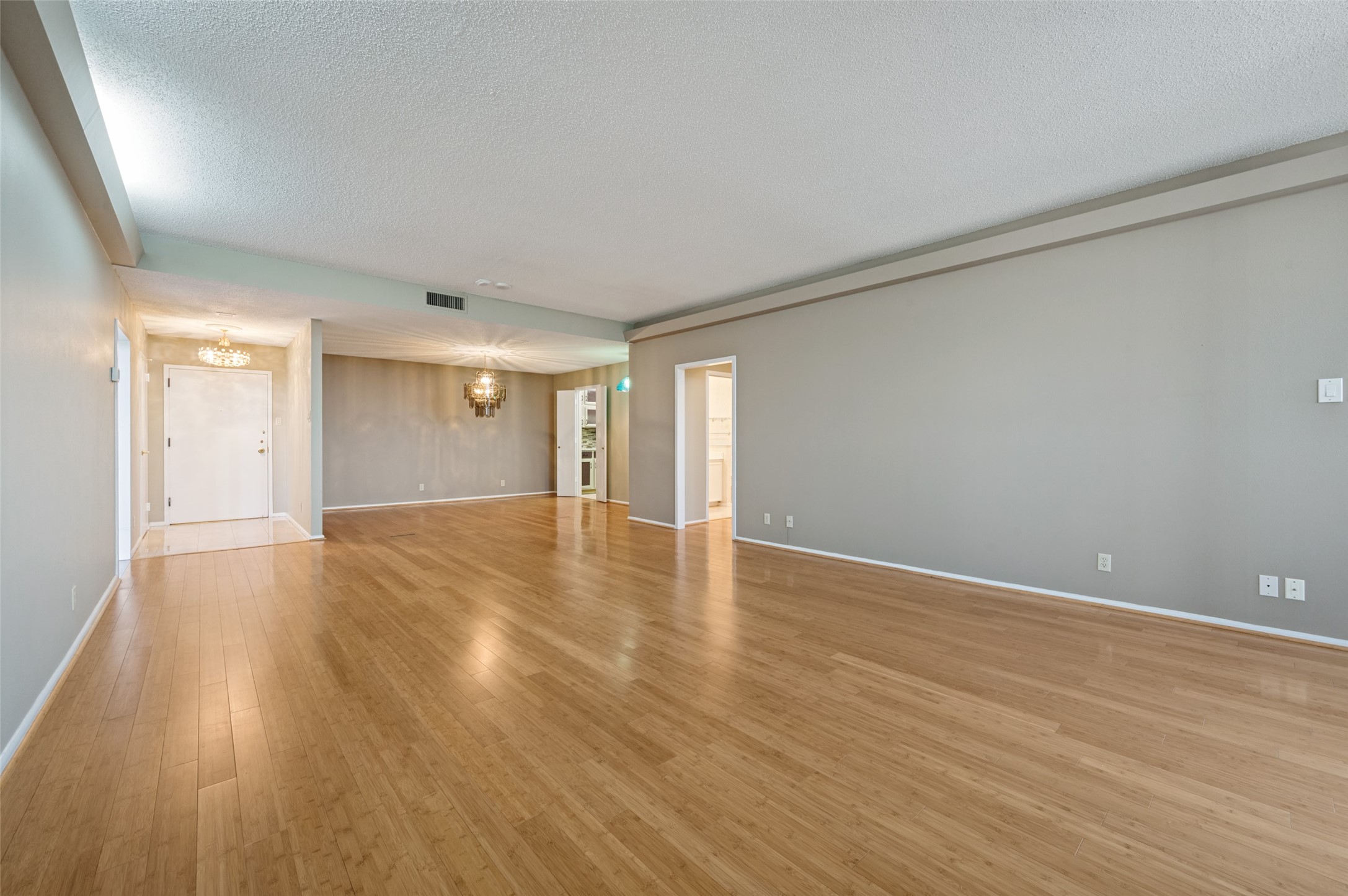 Empty living and dining area with hardwood floors, gray walls, and decorative chandeliers.
