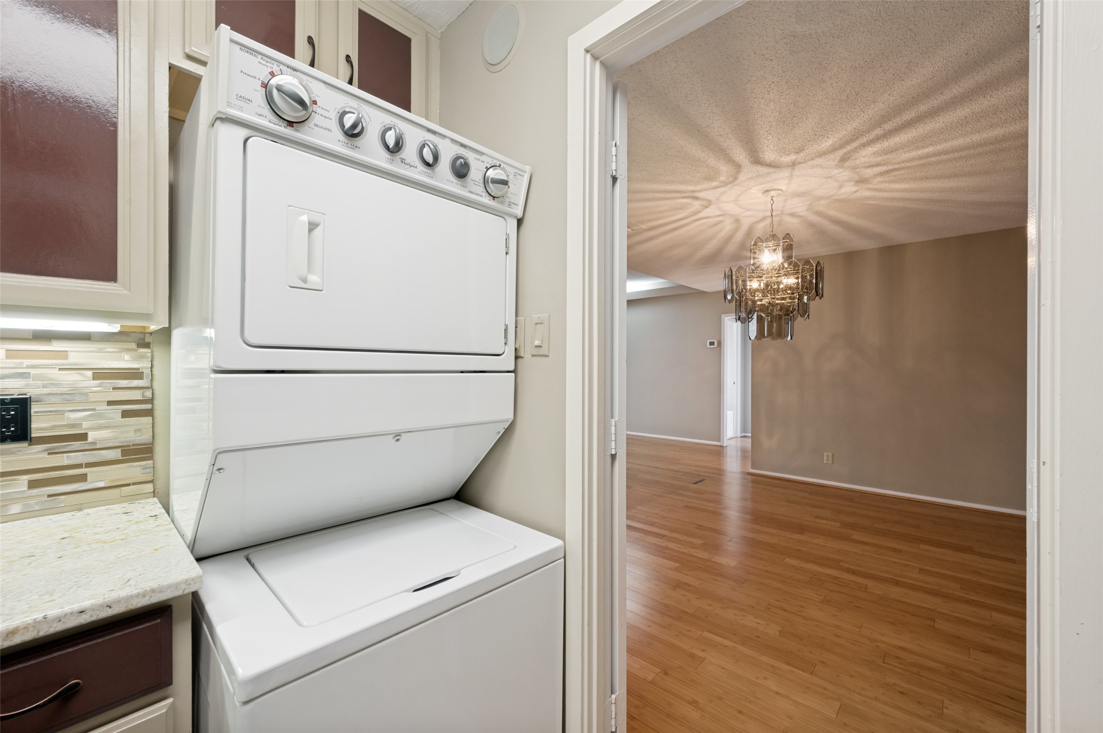 Laundry area with stacked washer and dryer beside a doorway to a hardwood-floored room with a crystal chandelier.