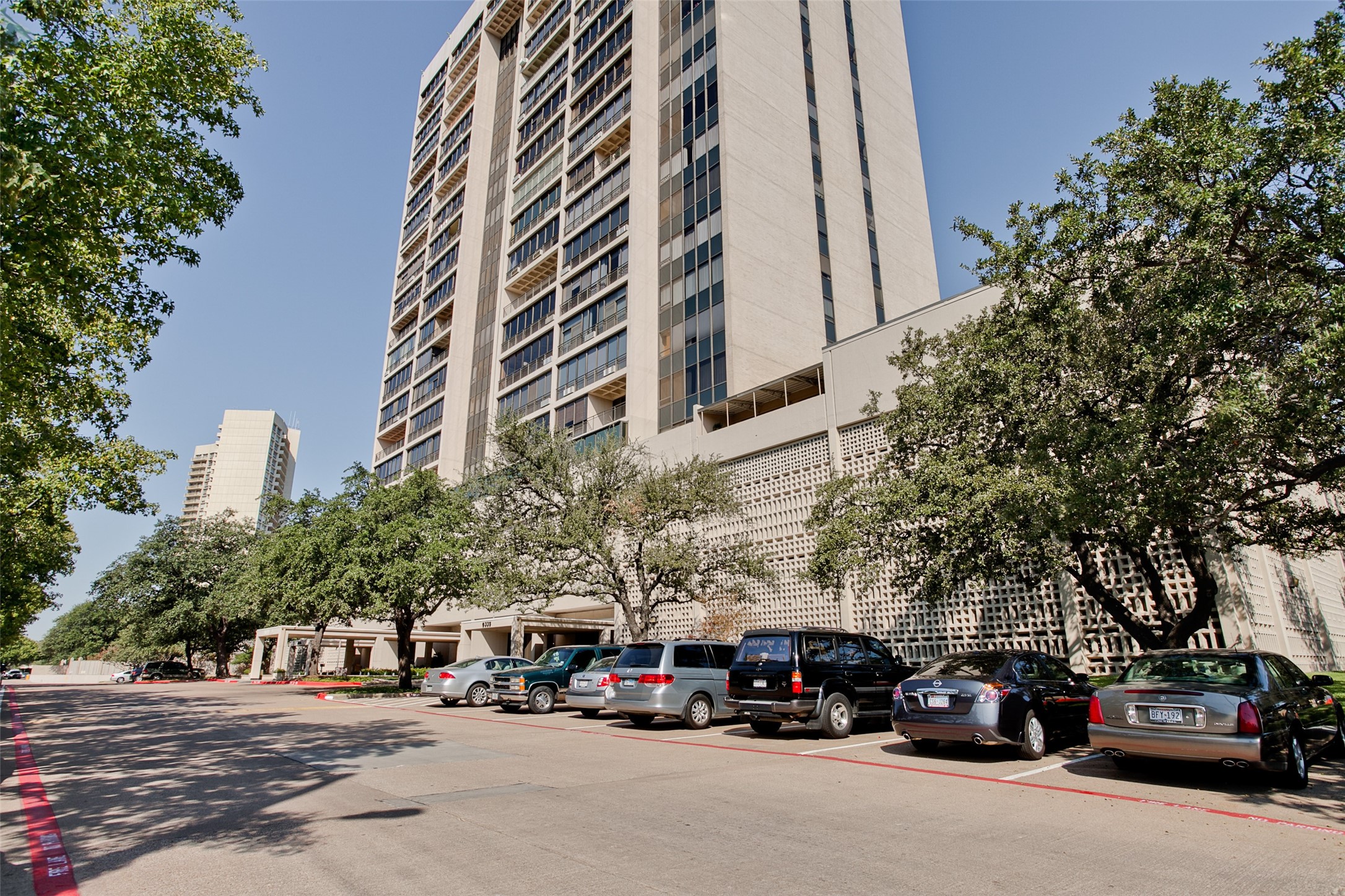 High-rise residential tower with balconies and a patterned facade, framed by trees and parked cars.