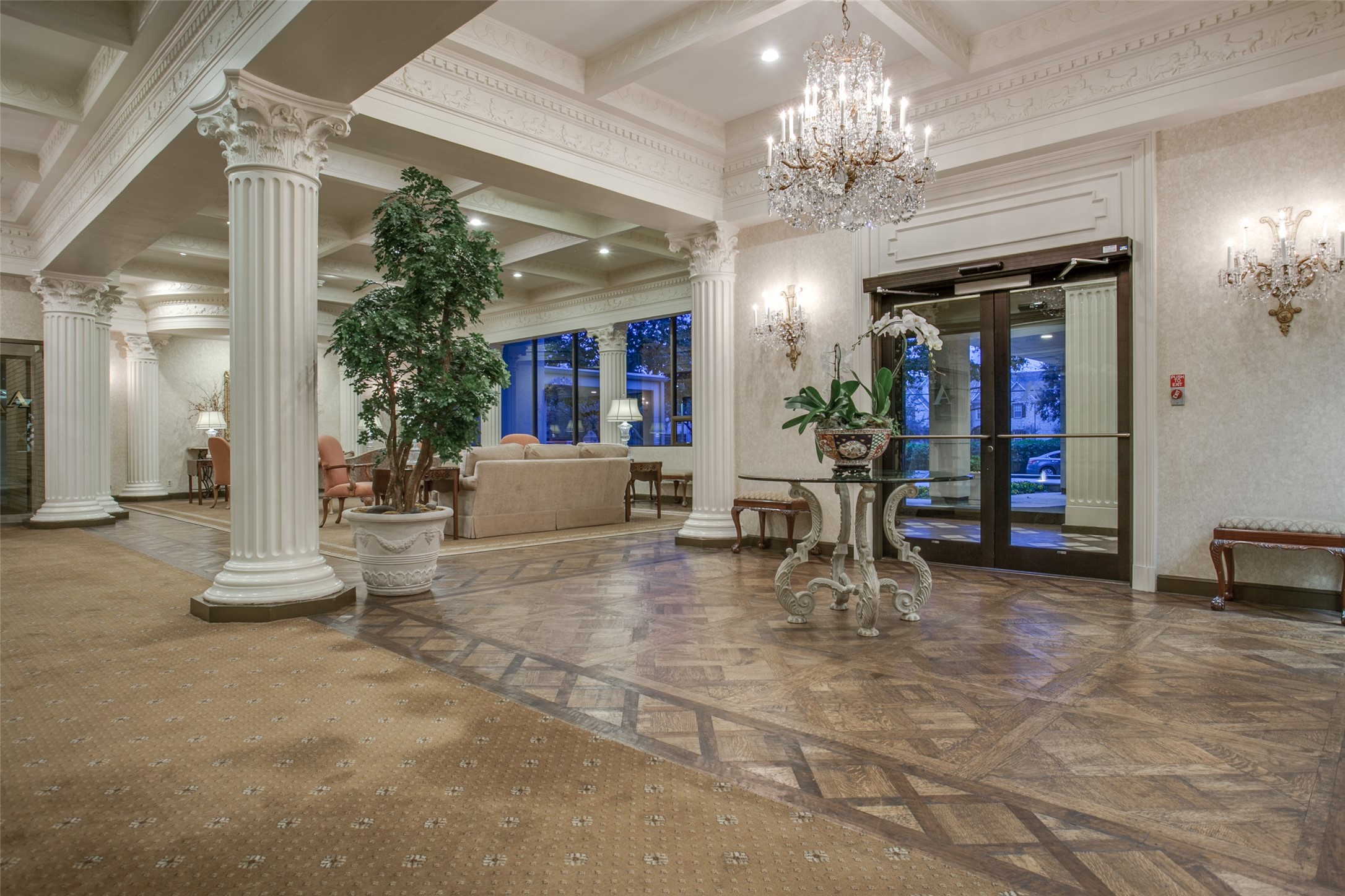 Residential lobby with fluted columns, a crystal chandelier, patterned wood flooring, and a potted tree.