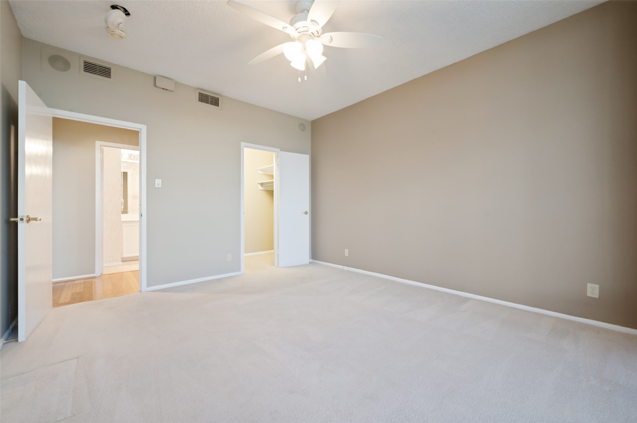 Empty carpeted bedroom with a ceiling fan and open doors to a bathroom and closet.