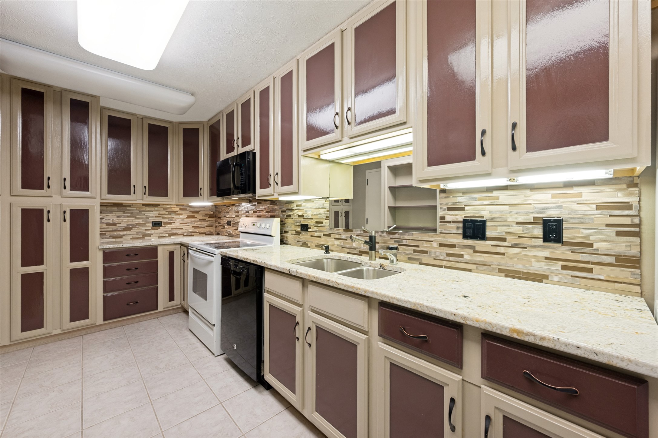 Kitchen with cream and burgundy cabinets, stone counters, a double sink, and multicolored tile backsplash.
